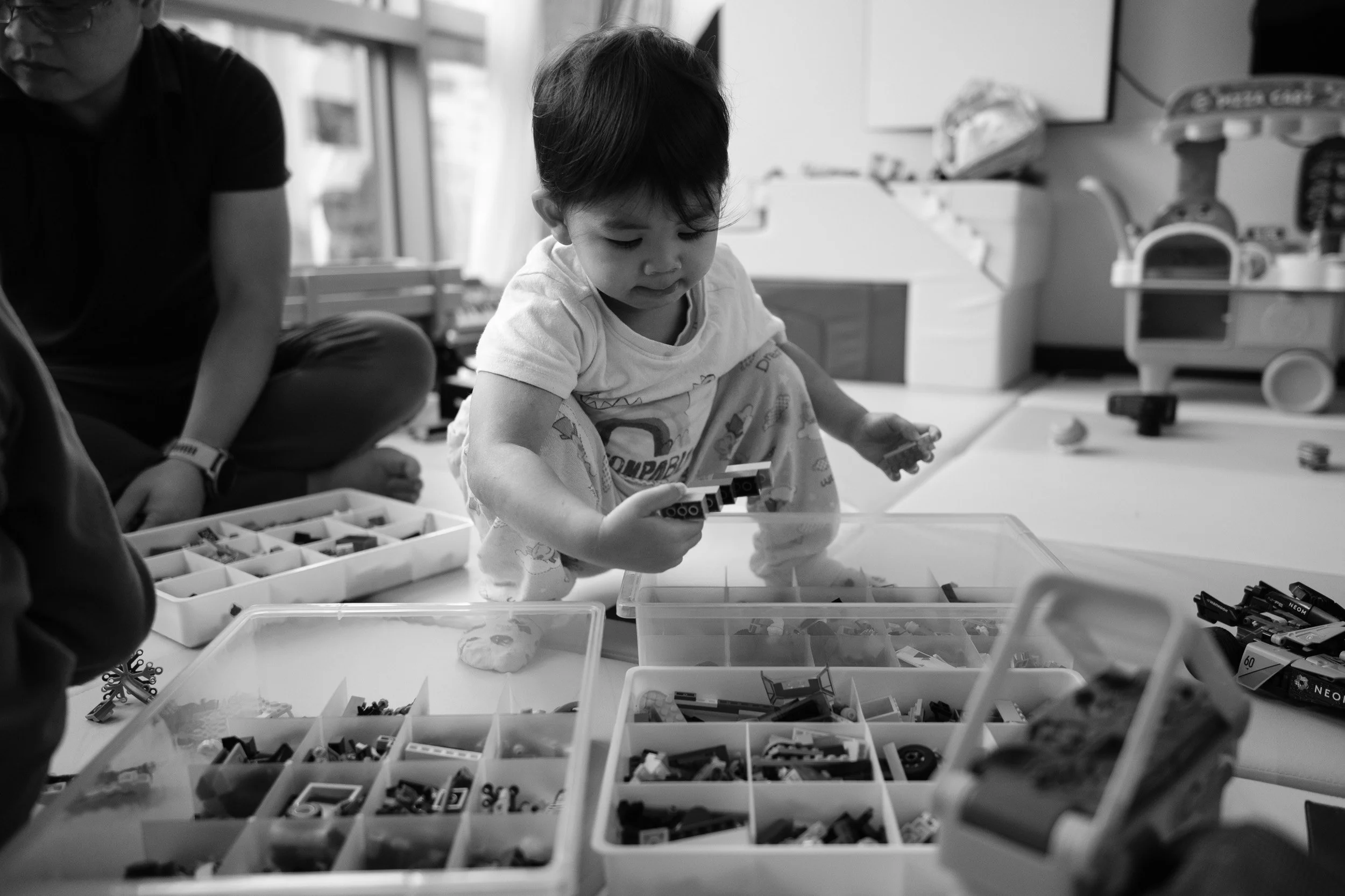 Black and white photo of a toddler sorting Lego bricks from organizer boxes on a playroom floor, documentary family photography Taipei