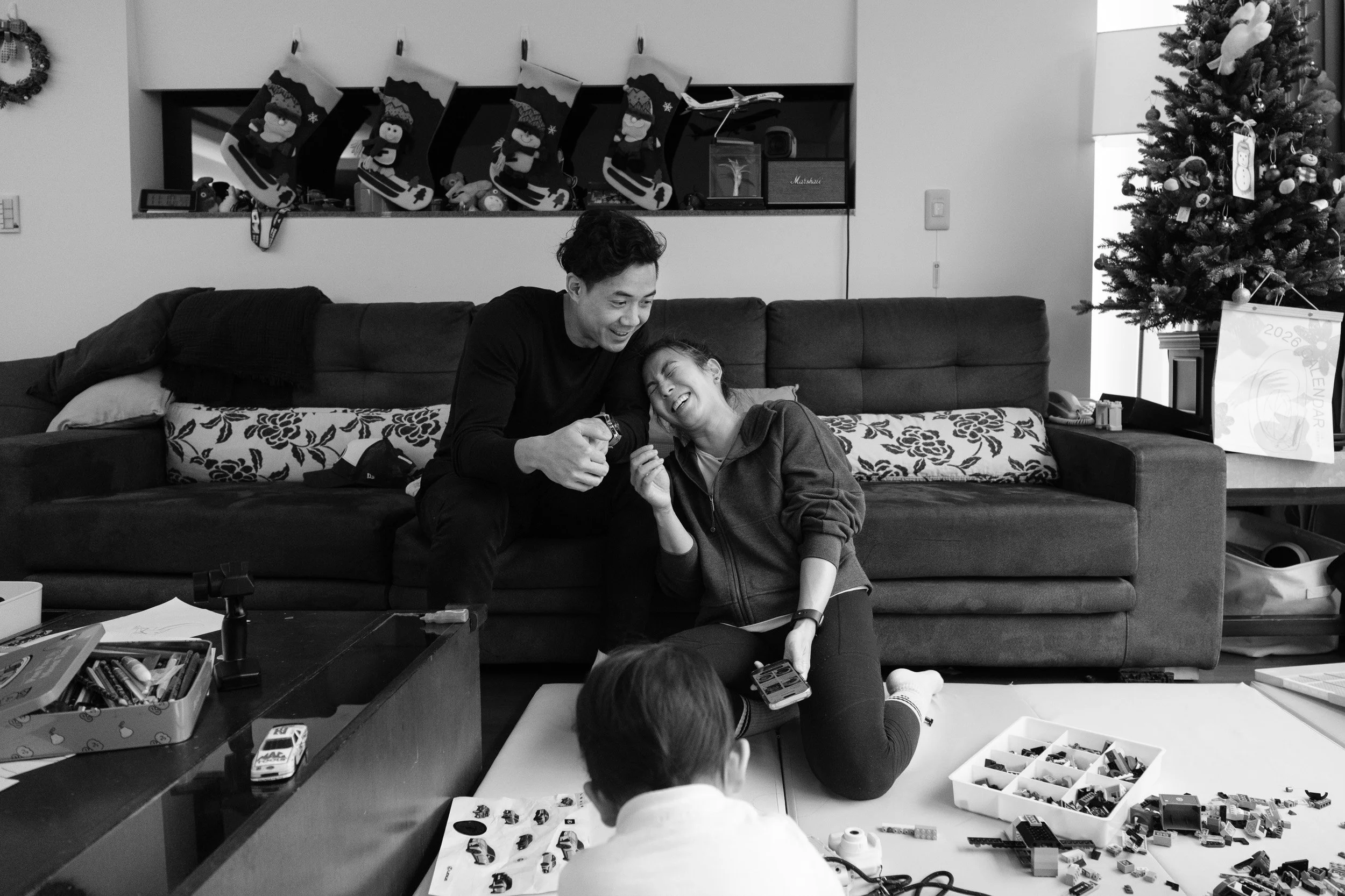 Black and white documentary family photography: a man and woman laugh together on a living room floor and sofa near a Christmas tree in Taipei
