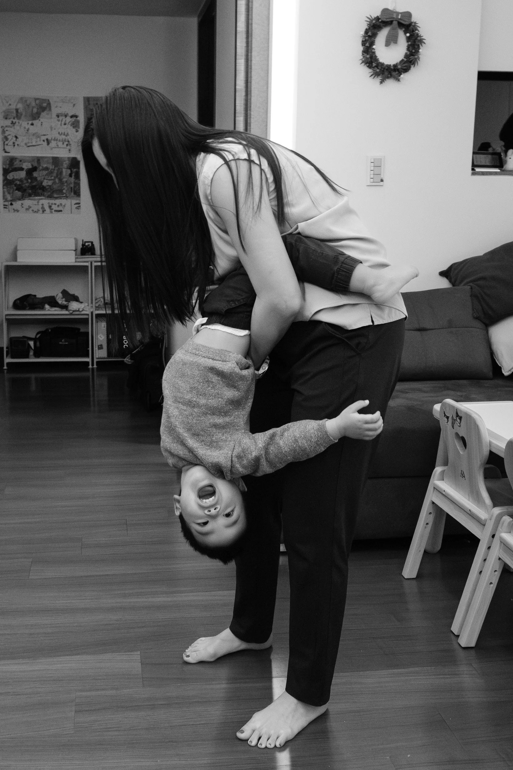 Black and white photo of a woman carrying a laughing toddler upside down in a Taipei living room, candid family photography