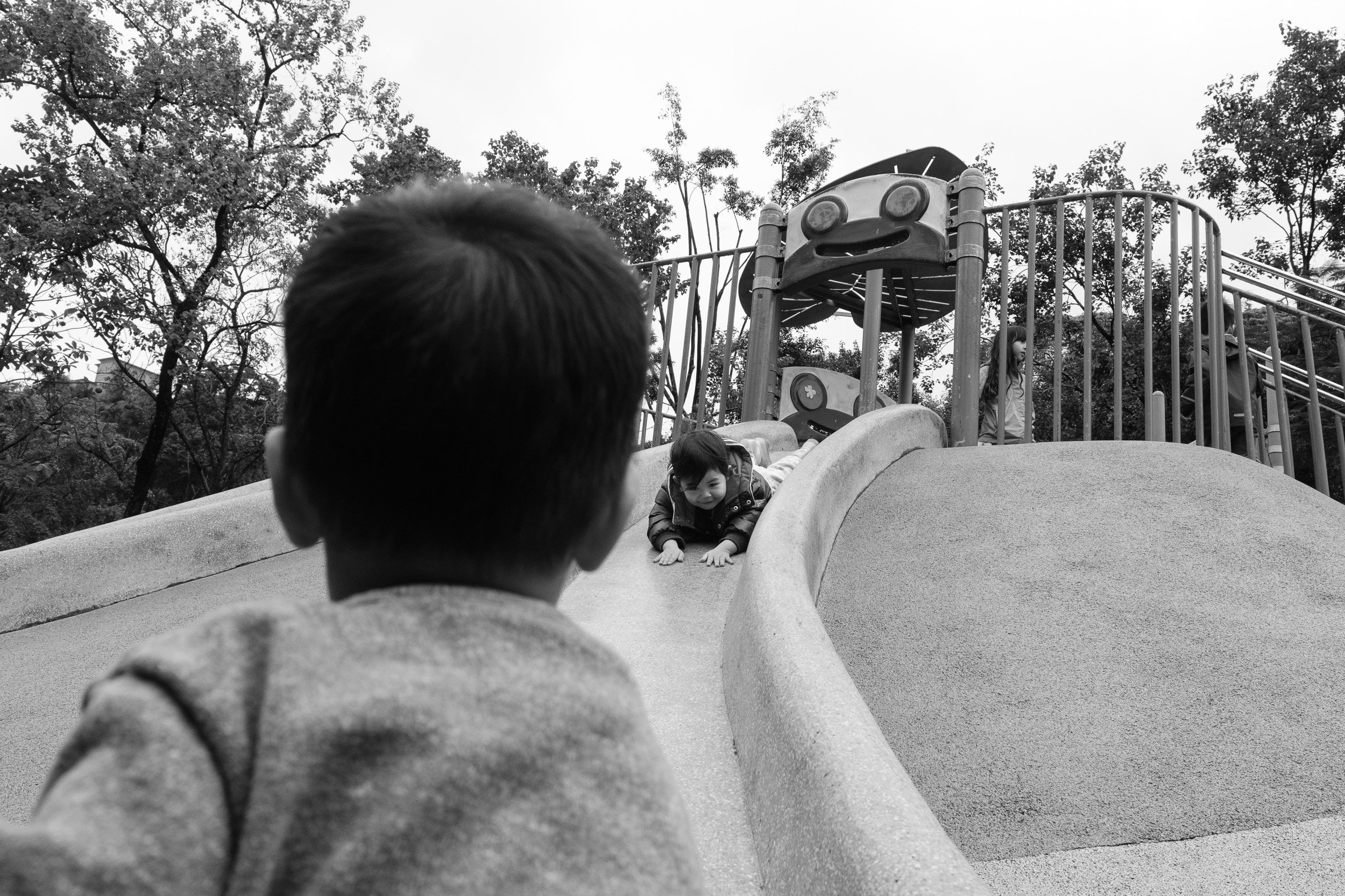 Black and white photo from behind a boy watching a toddler slide down a playground slide in Taipei, documentary family photographer
