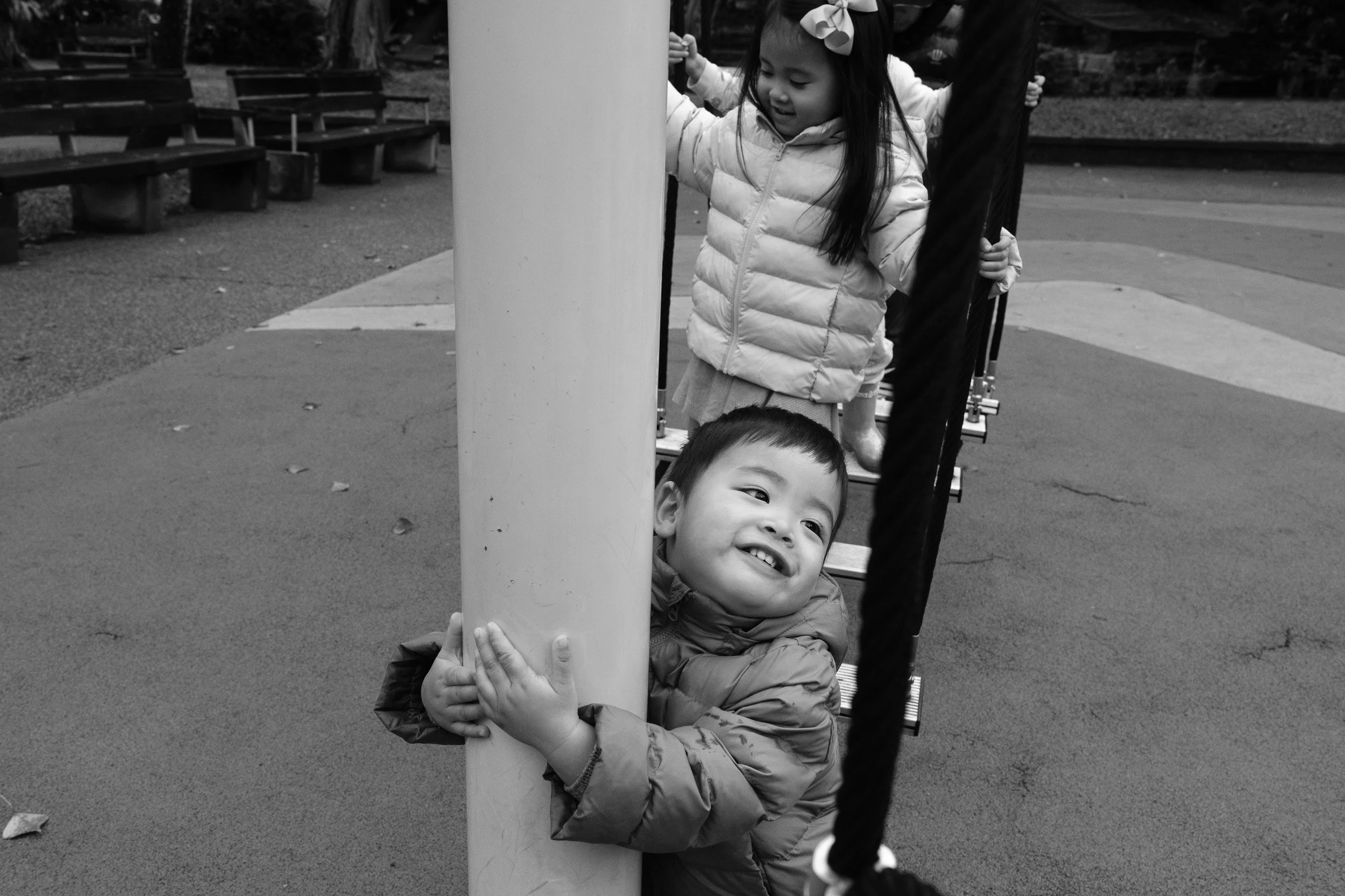 Black and white photo of a toddler hugging a playground pole and smiling while a girl stands on a rope bridge behind him, Taipei family photography