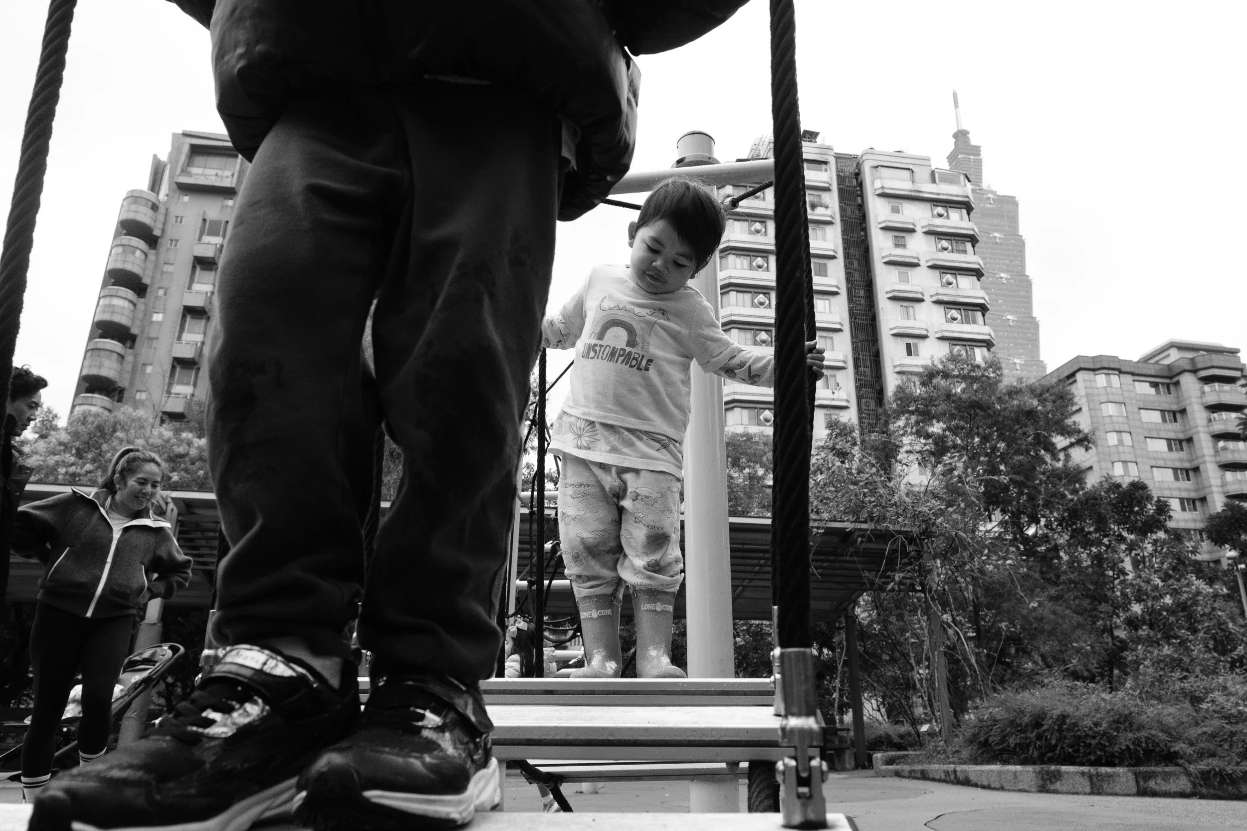 Black and white photo of a toddler crossing a rope-and-plank bridge at a Taipei playground, with an adult's legs in the foreground and Taipei city buildings behind