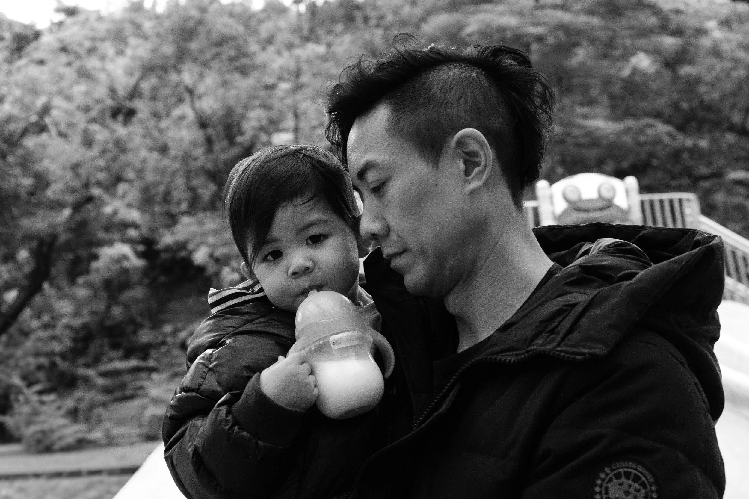 Black and white portrait of a man holding a toddler who drinks from a sippy cup at a Taipei playground, documentary family photography
