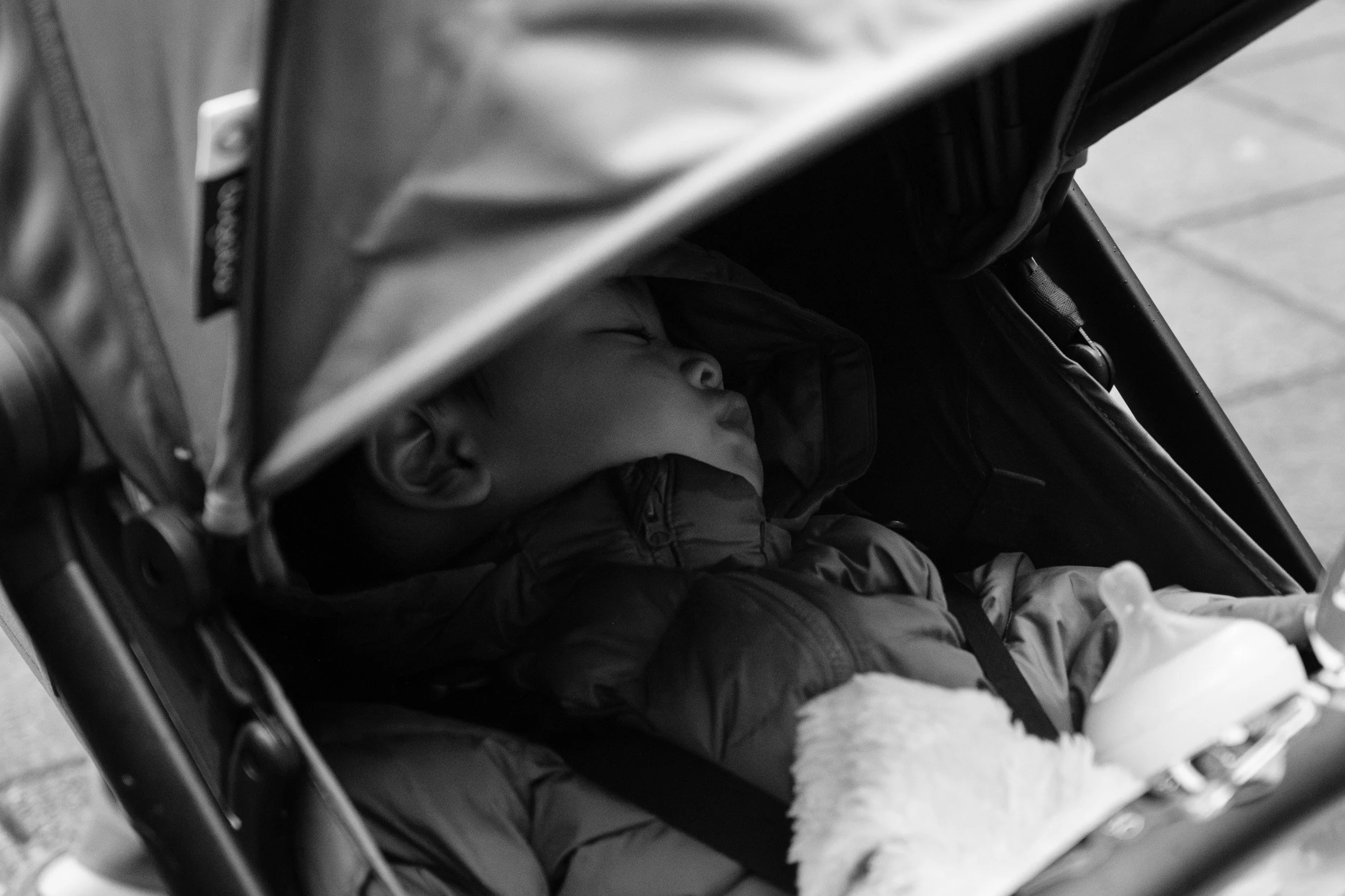 Black and white photo of a toddler asleep in a stroller in a puffy jacket, with a bottle beside him, documentary family photographer Taipei