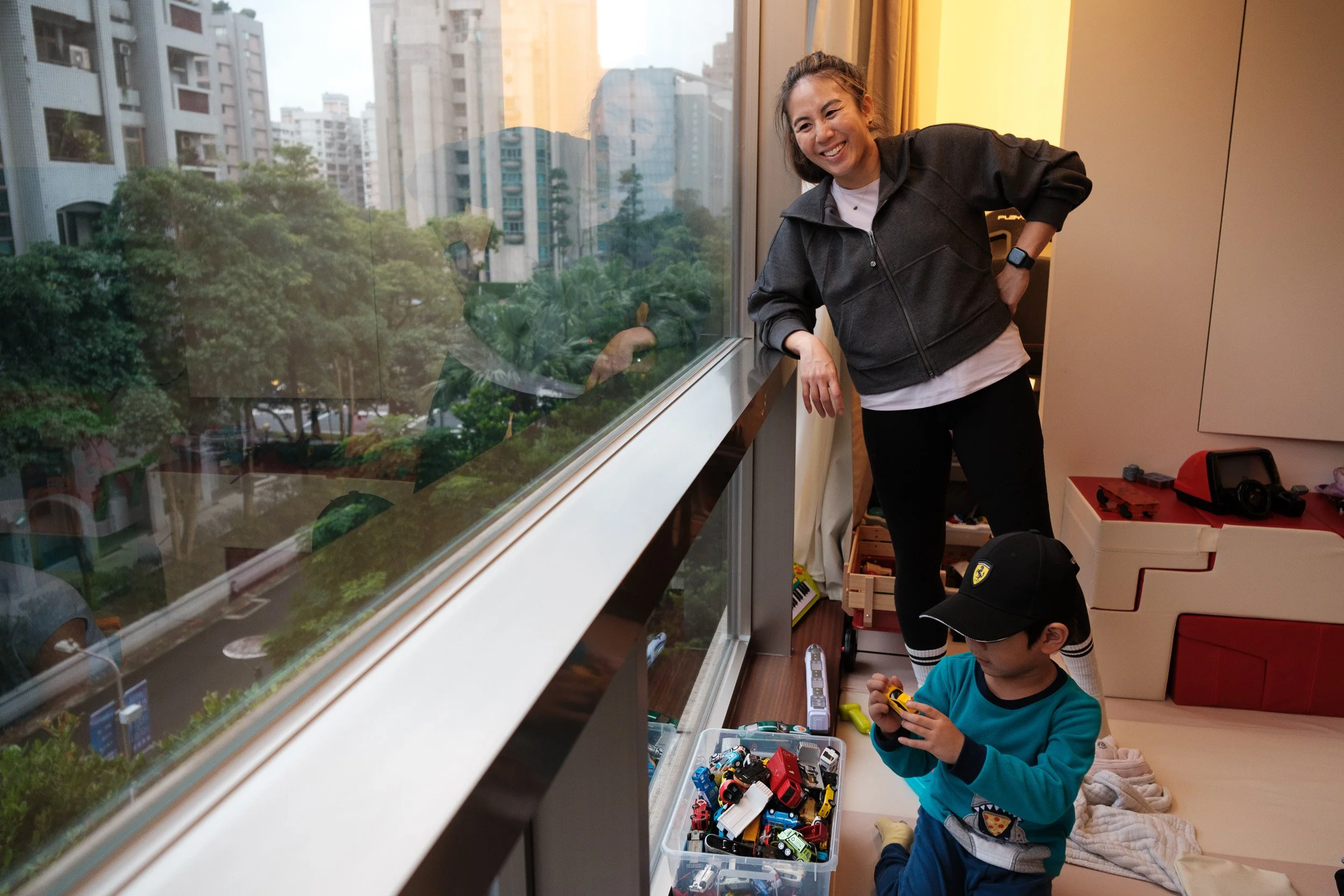 A woman leans on a window ledge and smiles at a boy in a Ferrari cap playing with toy cars in a Taipei apartment, documentary family photography
