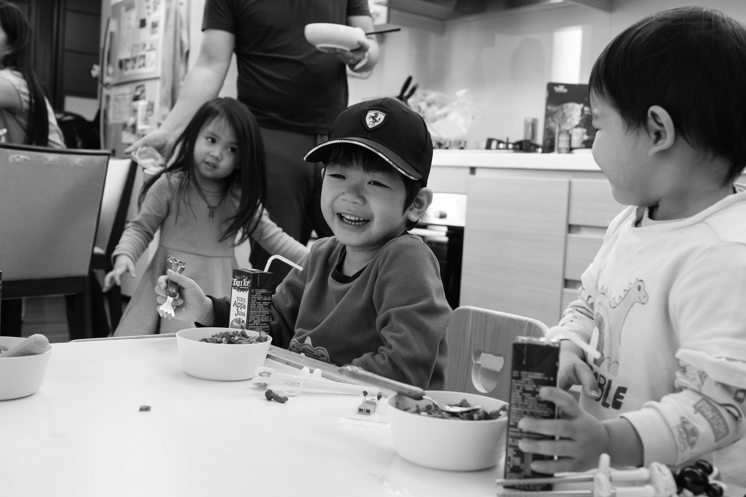 Black and white photo of a boy in a Ferrari cap laughing at a kitchen table with a toddler and a girl nearby, Taipei family photographer