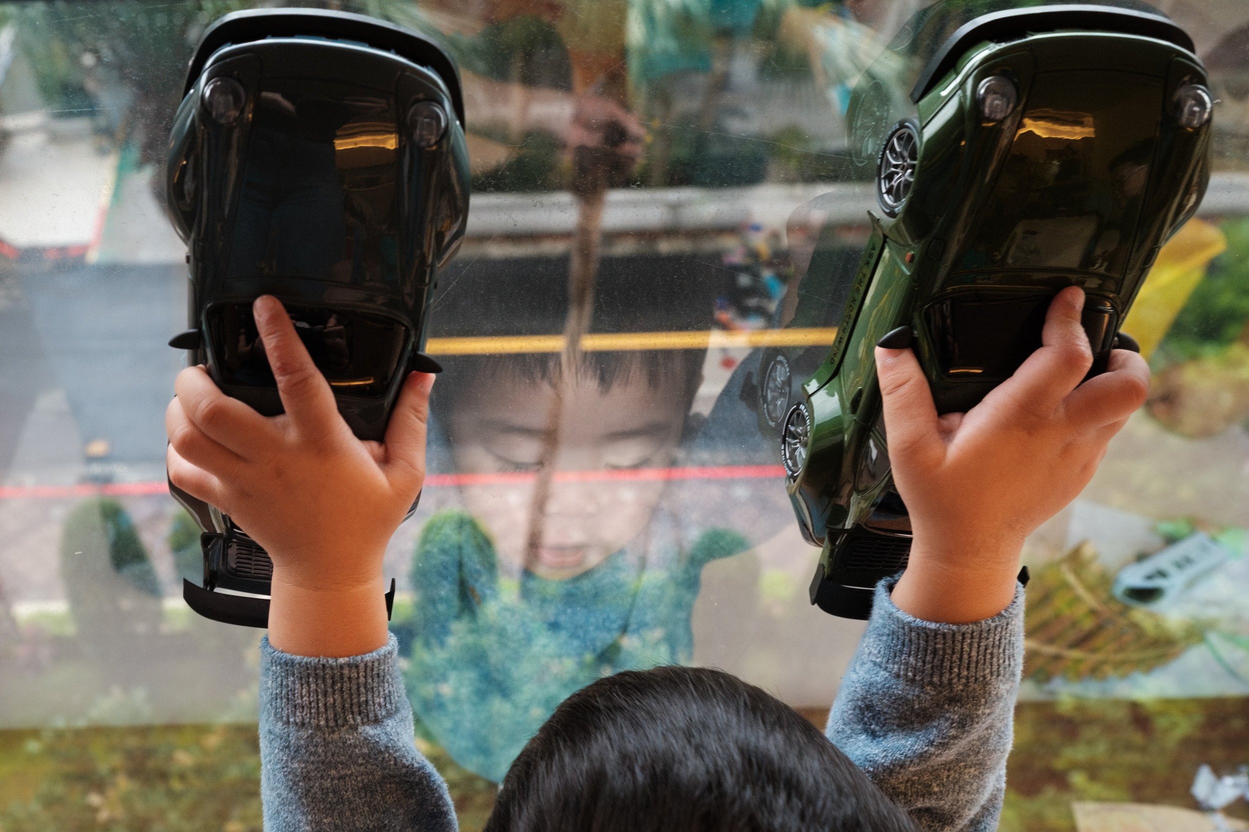 A toddler holds two toy cars up against a window, documentary family photography in Taiwan