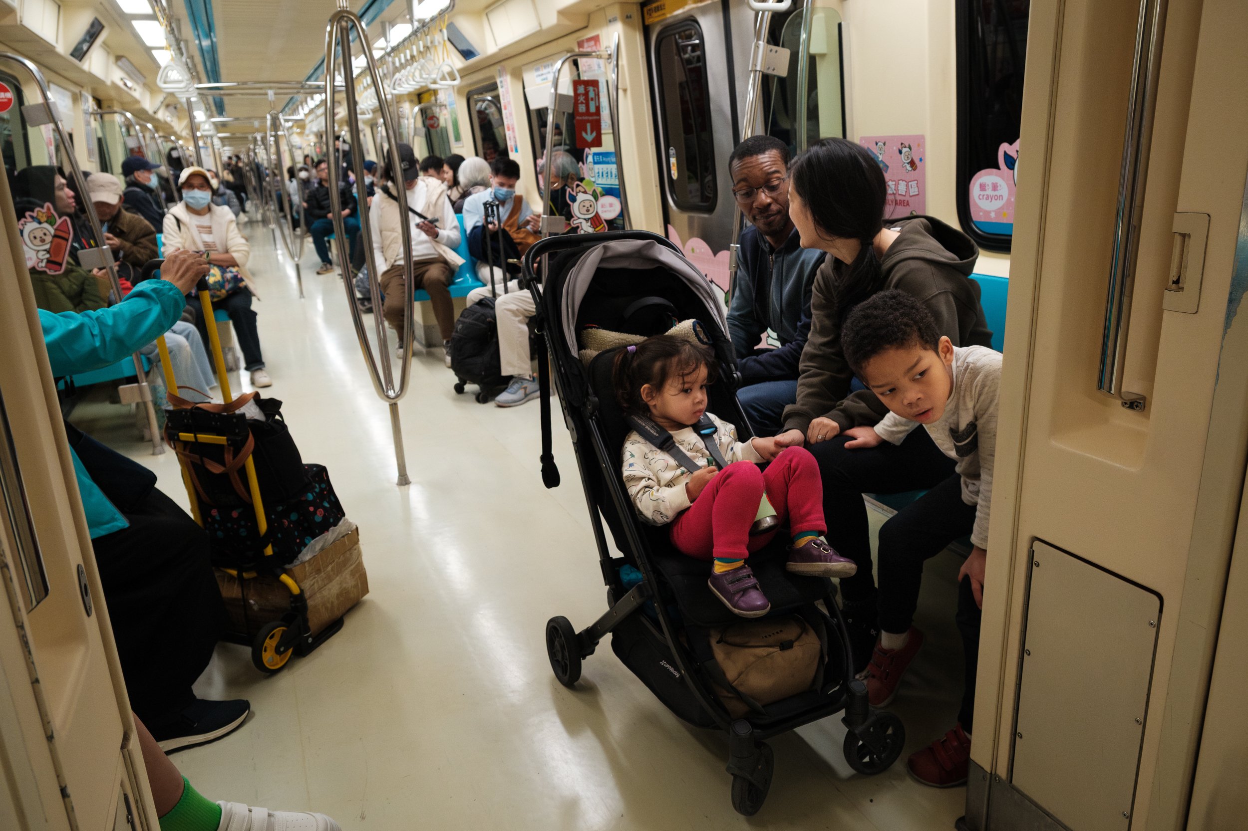 A color photo of a family of four sitting on the Taipei MRT. They have a stroller wth them and a crowd of people can be seen behind them.