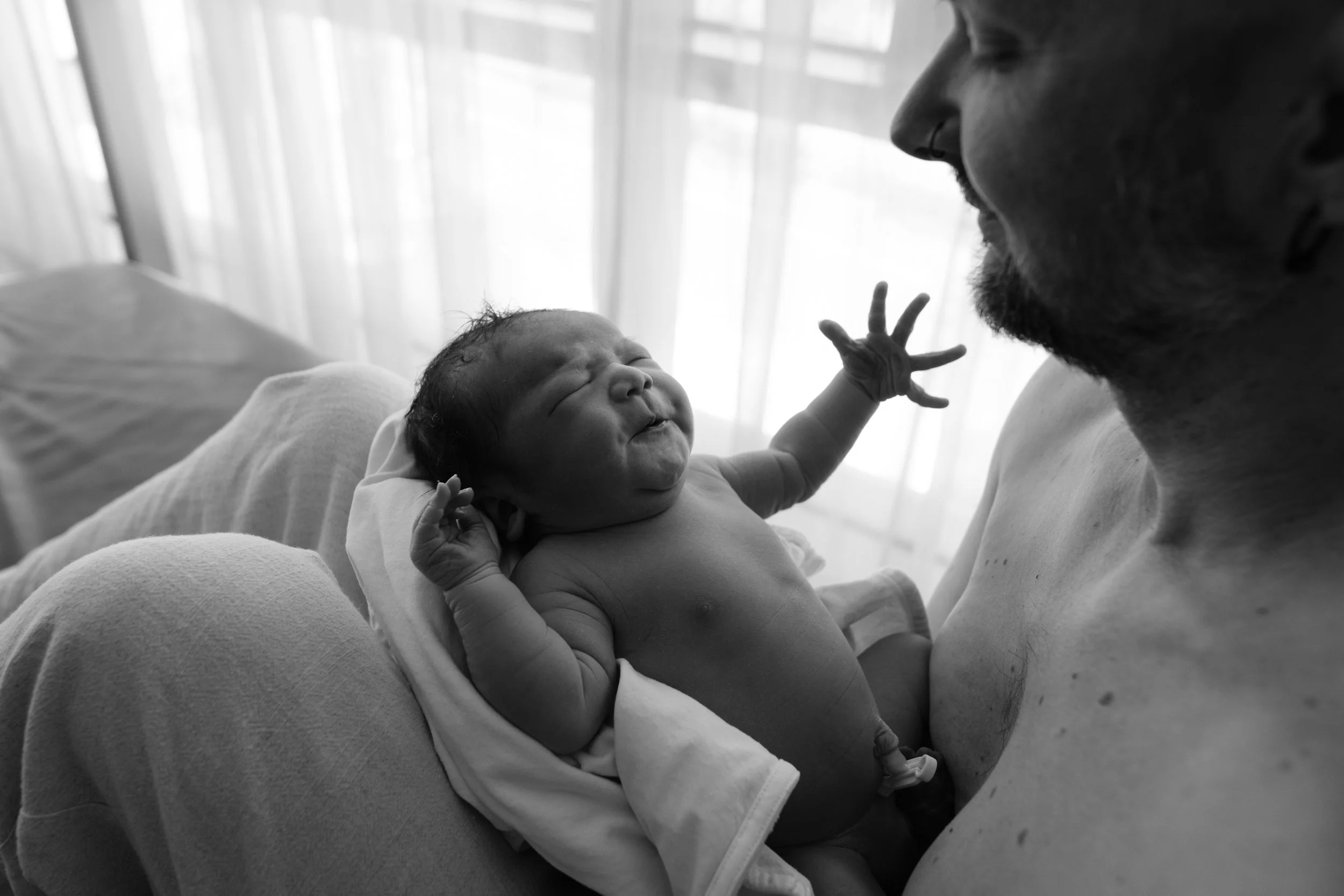 Black and white birth photography image of a father doing skin-to-skin with his newborn at Dianthus after a water birth, the baby's hand reaching out toward him.