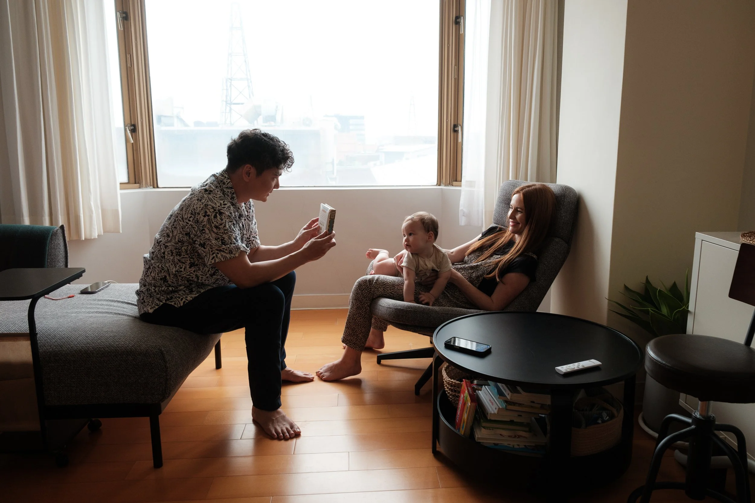 A woman is holding her baby in front of a big window in her living room in Taipei. A man is holding a book up for the baby to see.