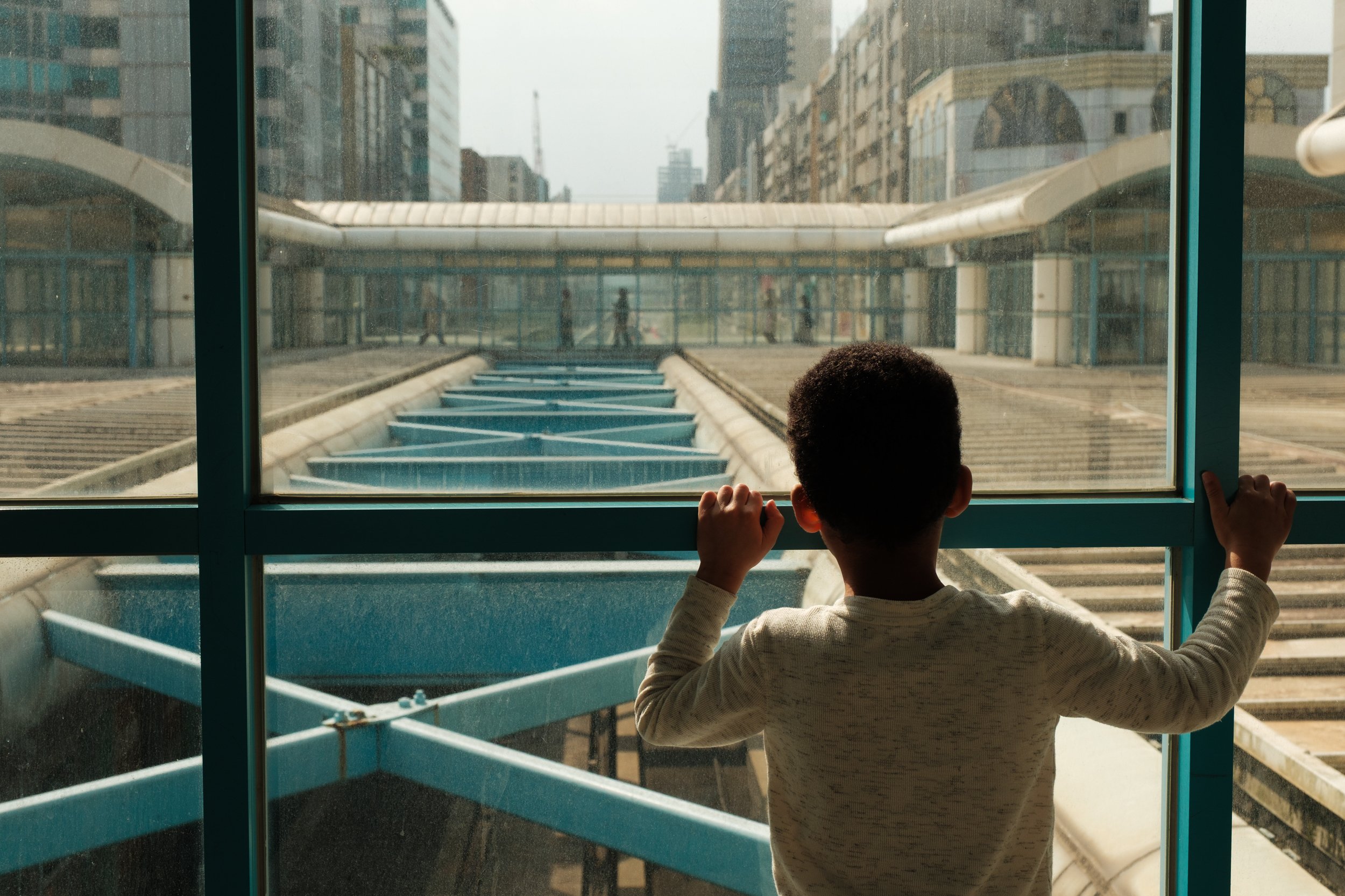 A color photo of a boy looking out a big window at Daan Station on the borwn line of the Taipei MRT. Other passengers can be seen on the opposite platform. 