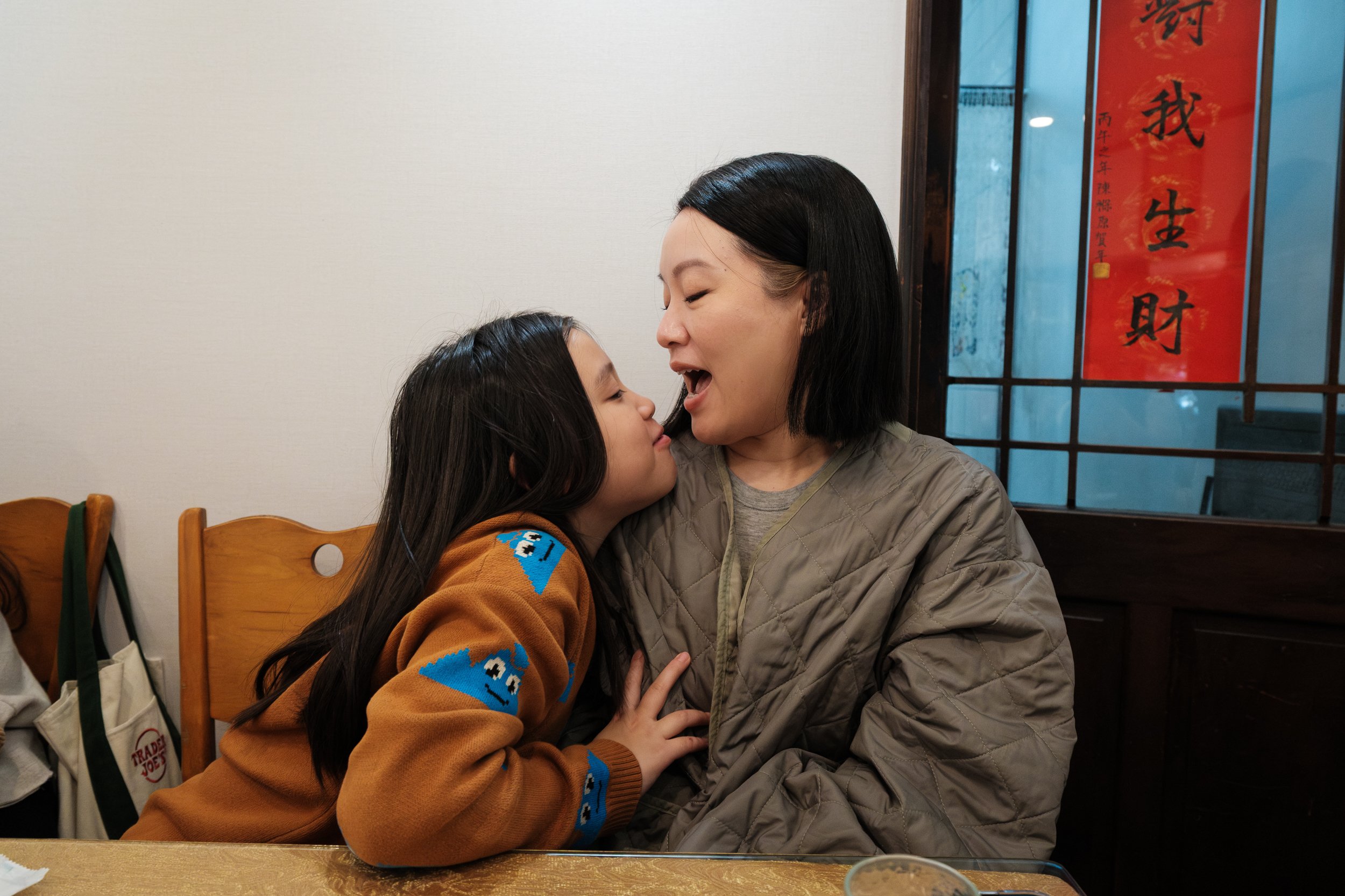 A woman and her young daughter are looking at each other and making funny faces at a restaurant in Tainan, Taiwan during a day in the life documentary family photography session.