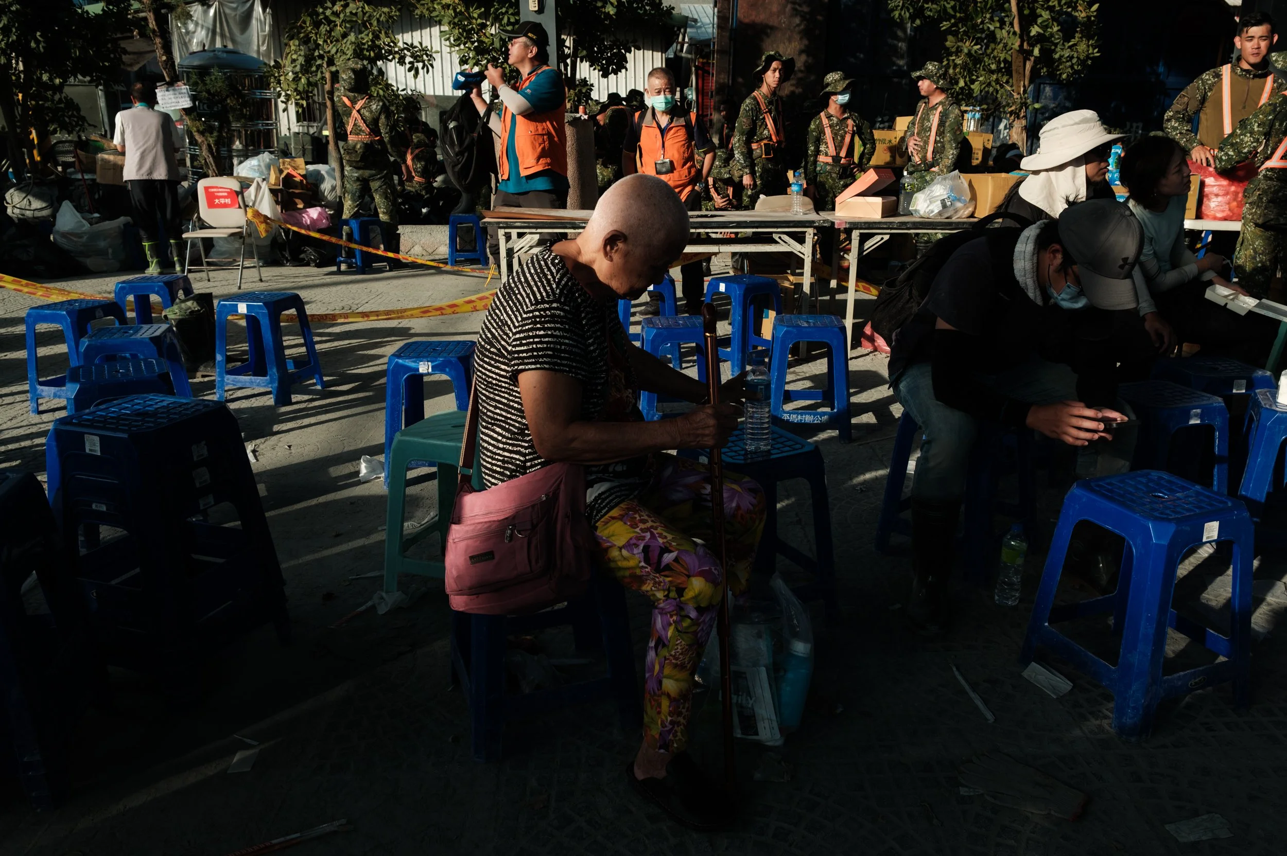 An old woman waits for aid at a community center in Guangfu. She is hunshed over and military personnel can be seen behind her.