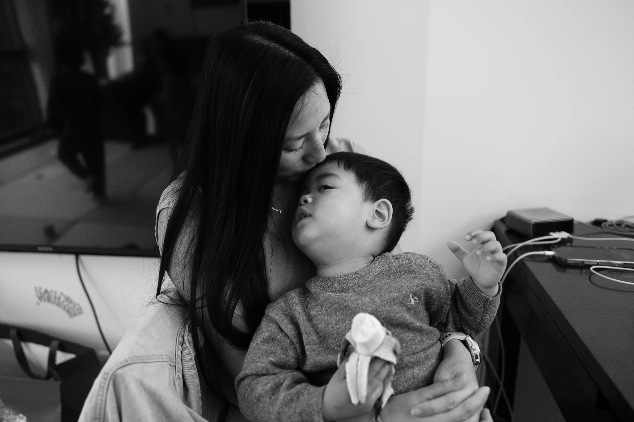 A woman is kissing the top of her toddler son's head as he eats a banana in their Taipei apartment.