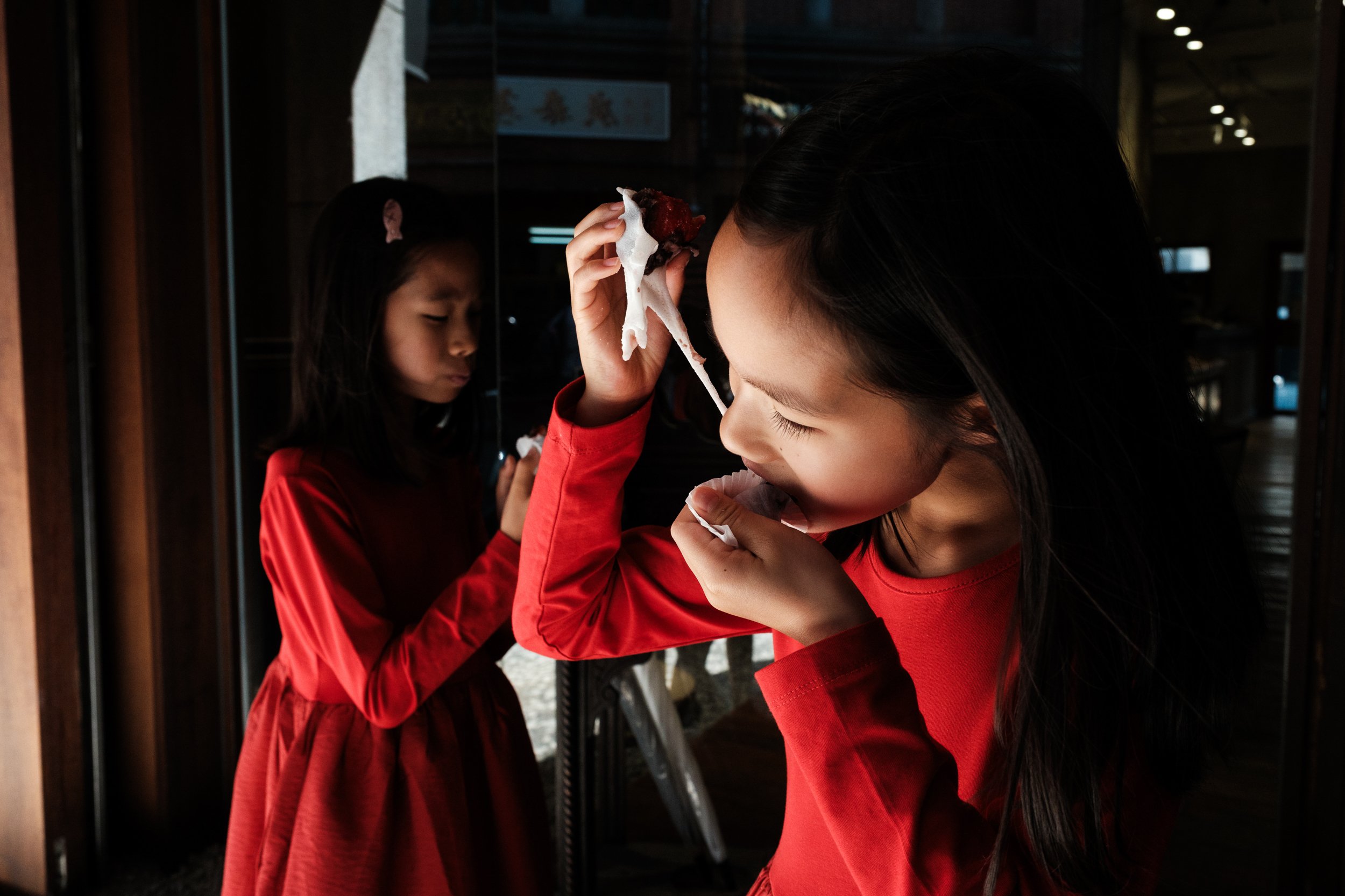 Two girls in matching red dresses eat mochi on Dihua Street in Taipei. One girl is stretching the mochi from her mouth. Photographed during an outdoor family photoshoot.