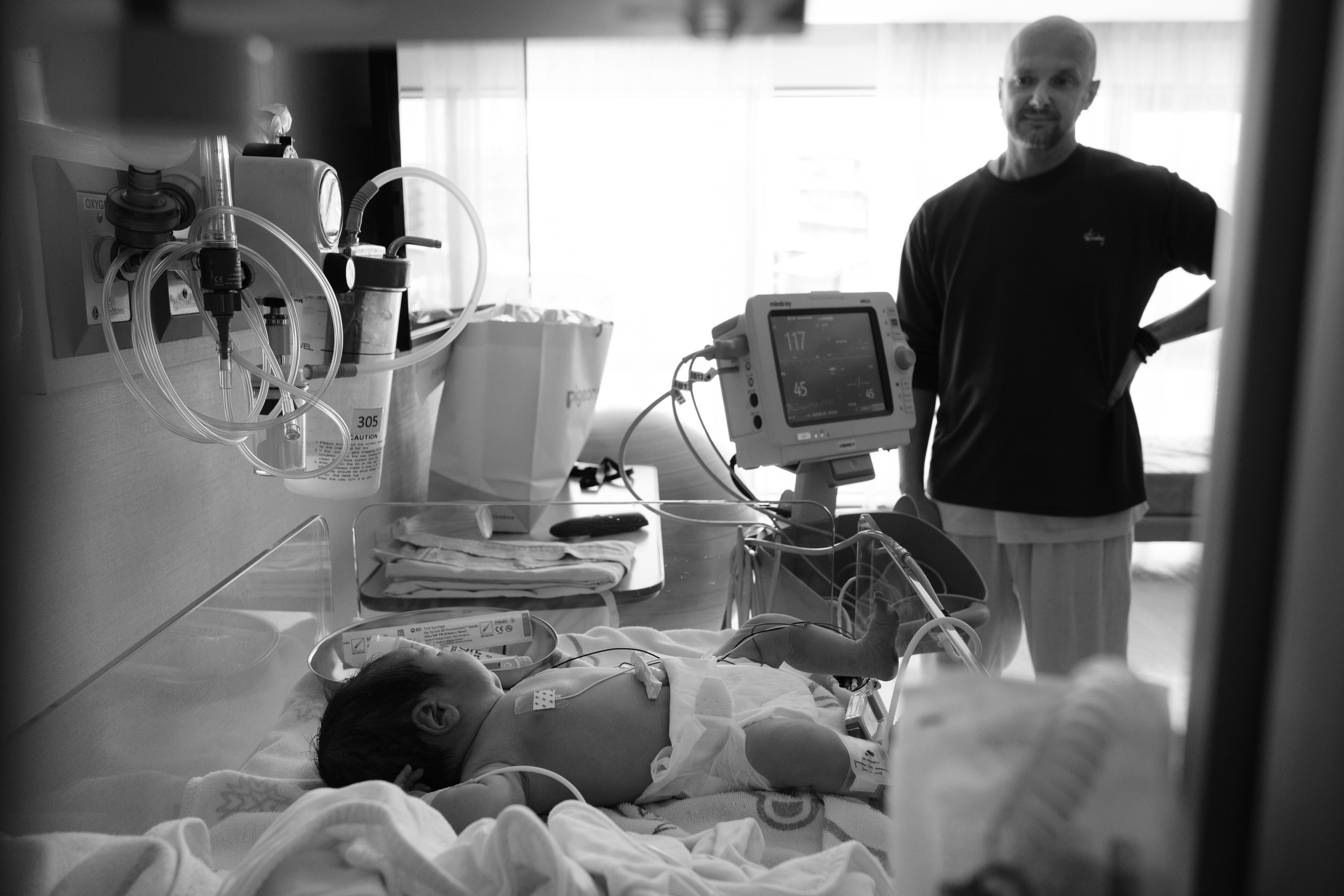 Black and white birth photography image of a newborn resting in a hospital bassinet with monitoring equipment at Dianthus, the father standing in the background.