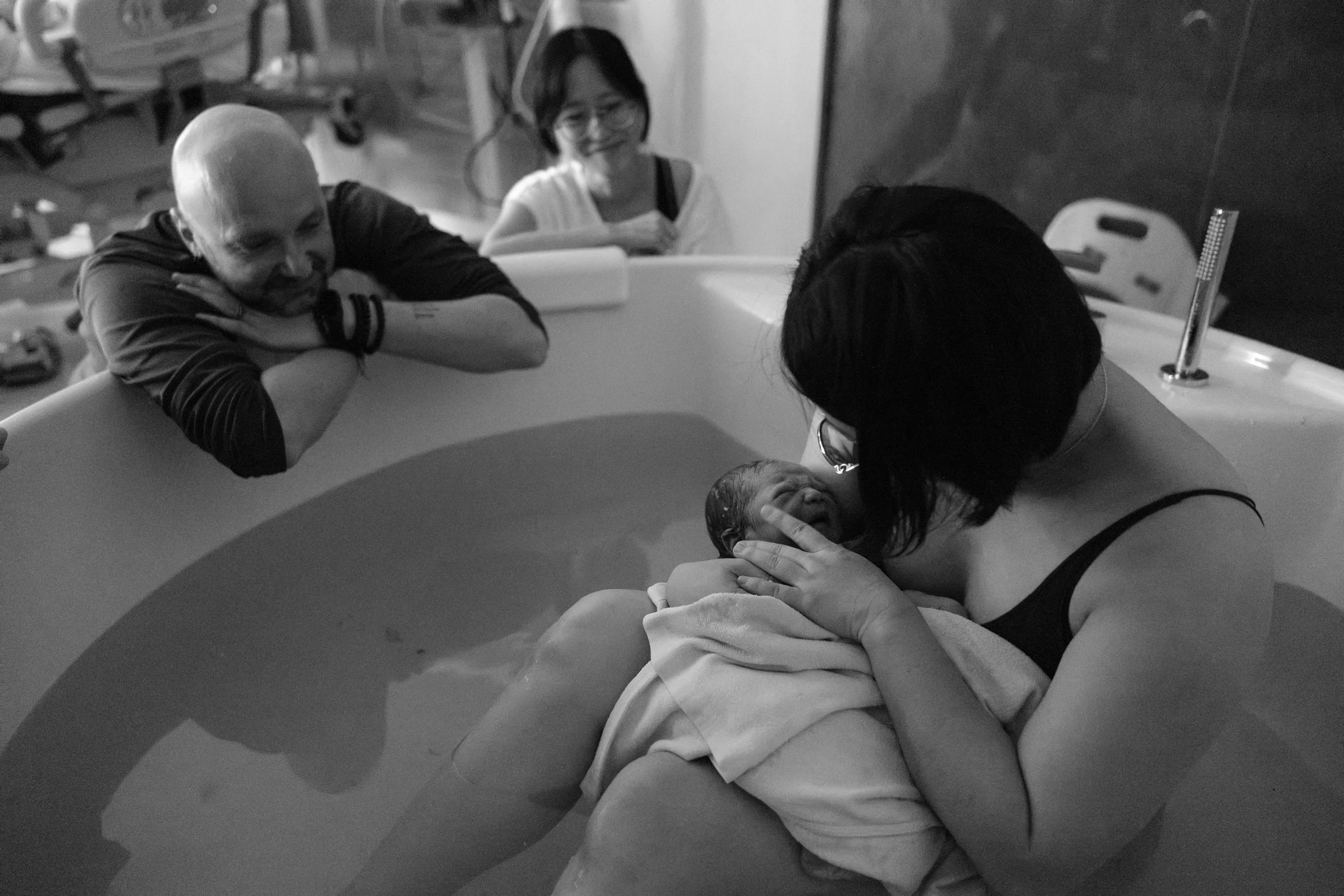 Black and white birth photography image of a mother holding her newborn in the birth pool moments after a water birth at Dianthus, with her partner and a nurse looking on.