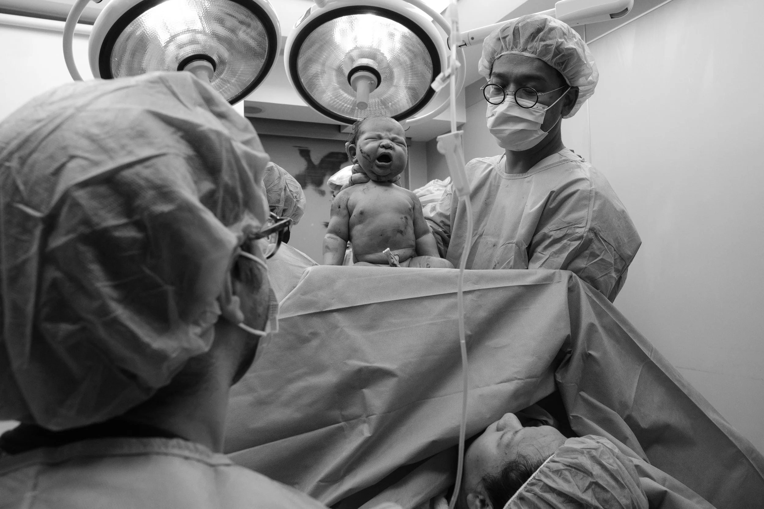 Black and white birth photography image of a newborn being held up under surgical lights immediately after a c-section at Dianthus, a doctor standing nearby.