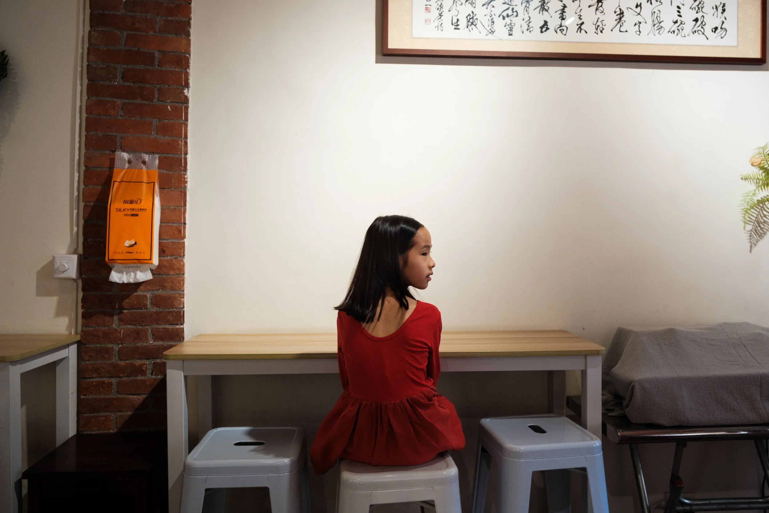 A young girl in a red dress is sitting on a plastic white stool in a shaved ice shop on Dihua Street in Taipei. Chinese caligraphy is hanging on the wall behind her.