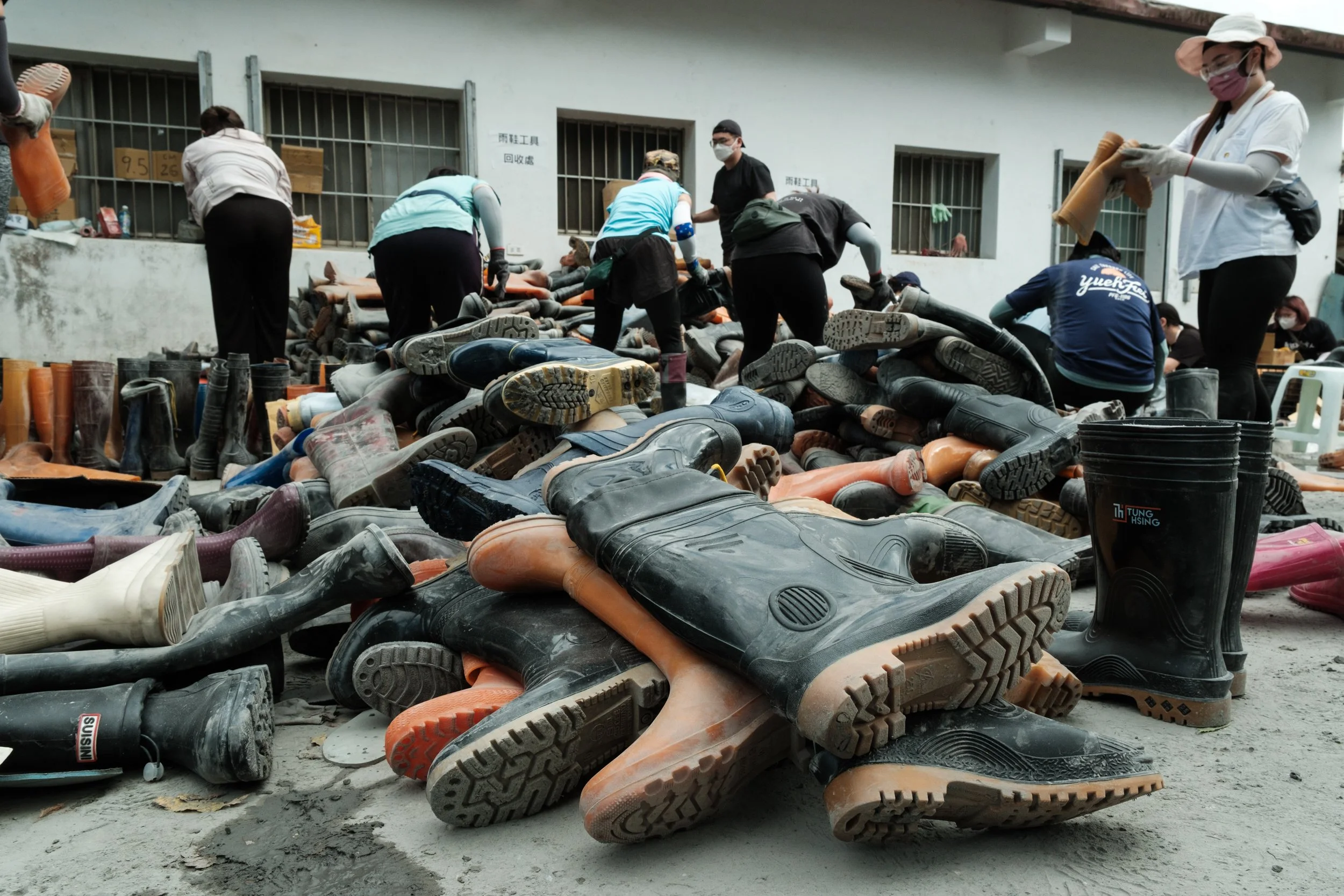 A pile of dirty rubber work boots is in the foreground and Guangfu flood recovery volunteers can be seen in the background cleaning.