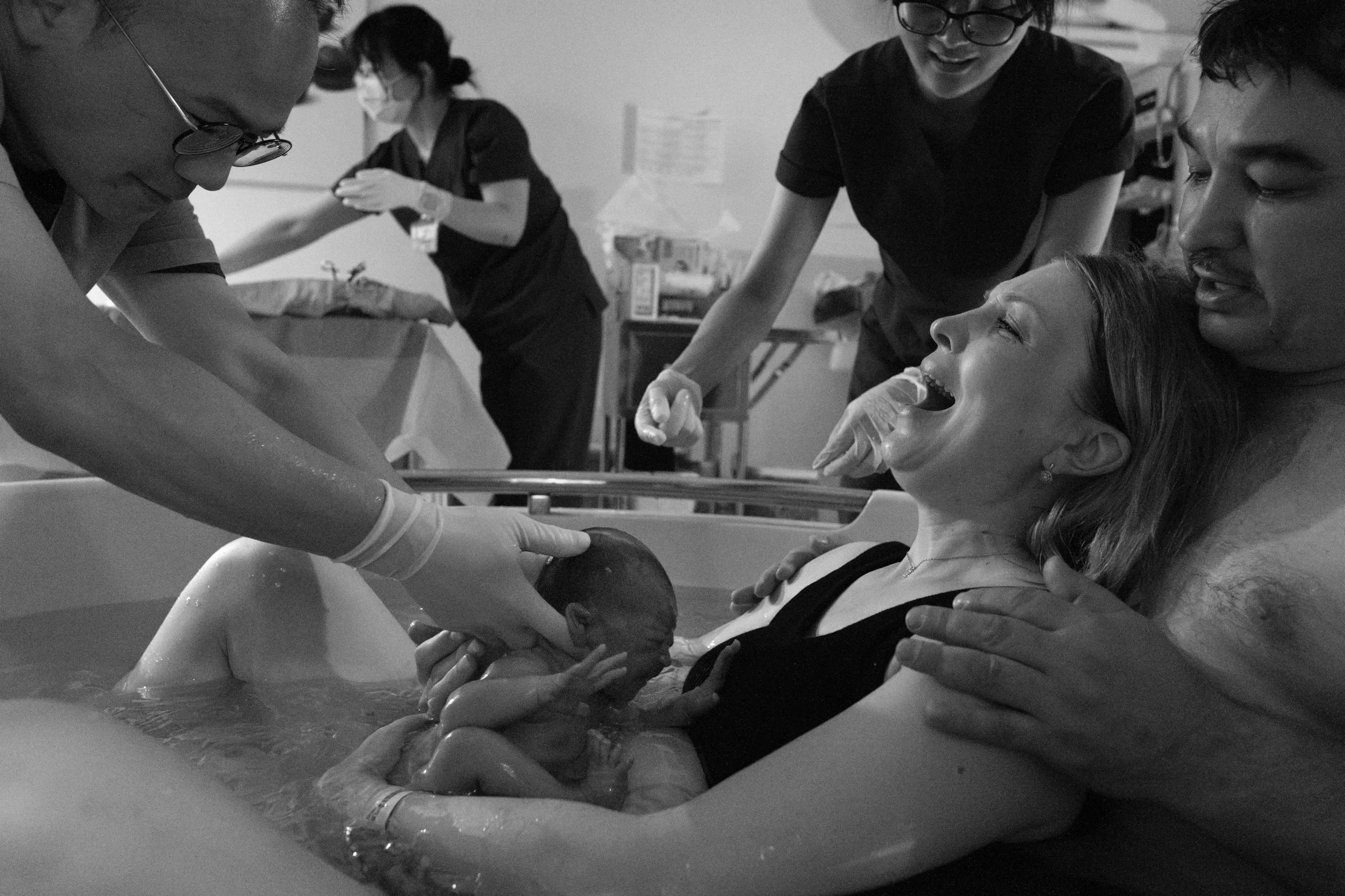 A black and white image of a newborn baby being born in a water birth tub at Dianthus in Neihu. A nurse is lifting the baby out of the water and handing her to the mom, who is smiling.