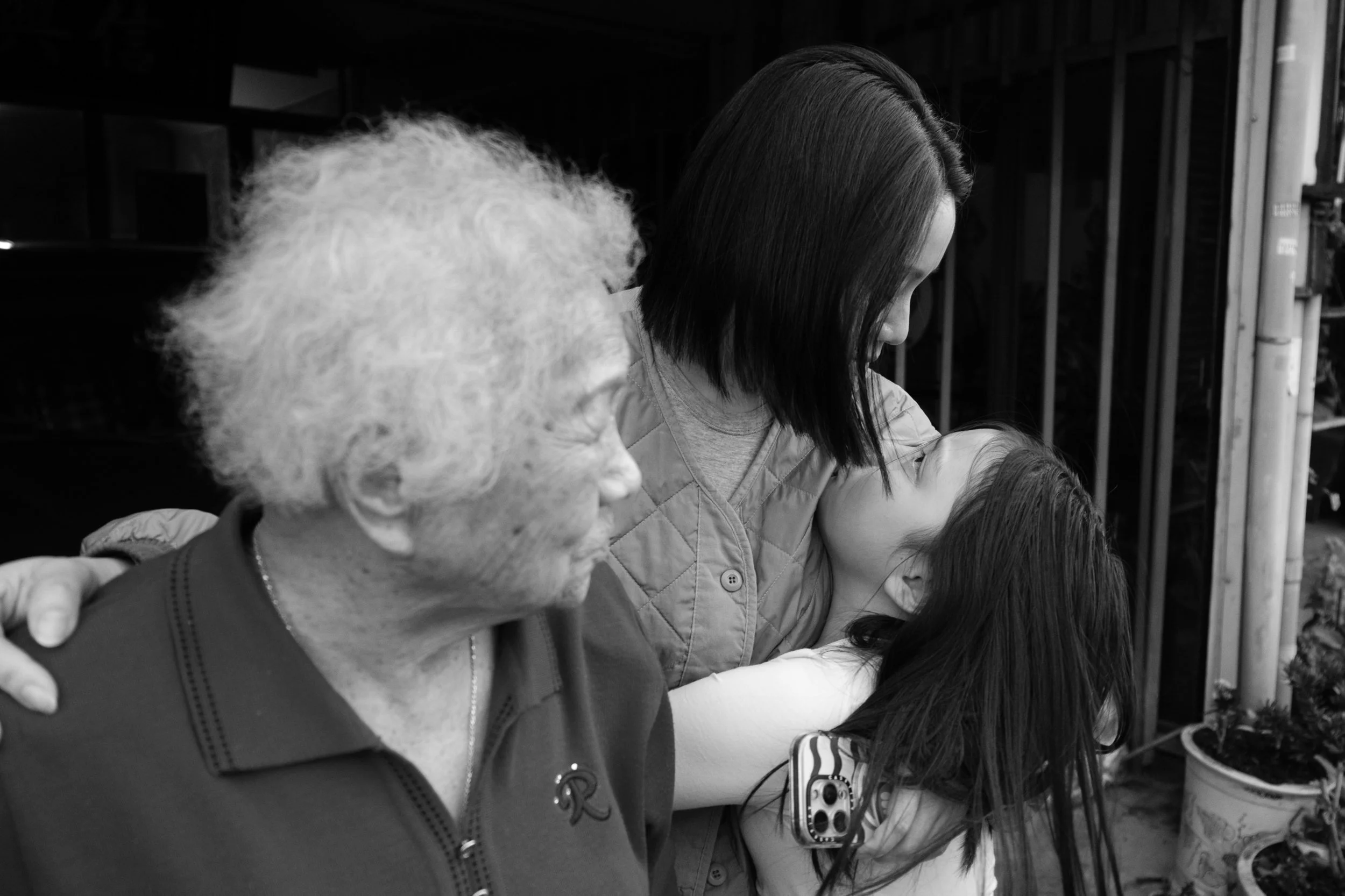 A woman is looking down into her daughter's face as she hugs her. An older woman is standing in front of them. Photographed during a family session in Tainan.