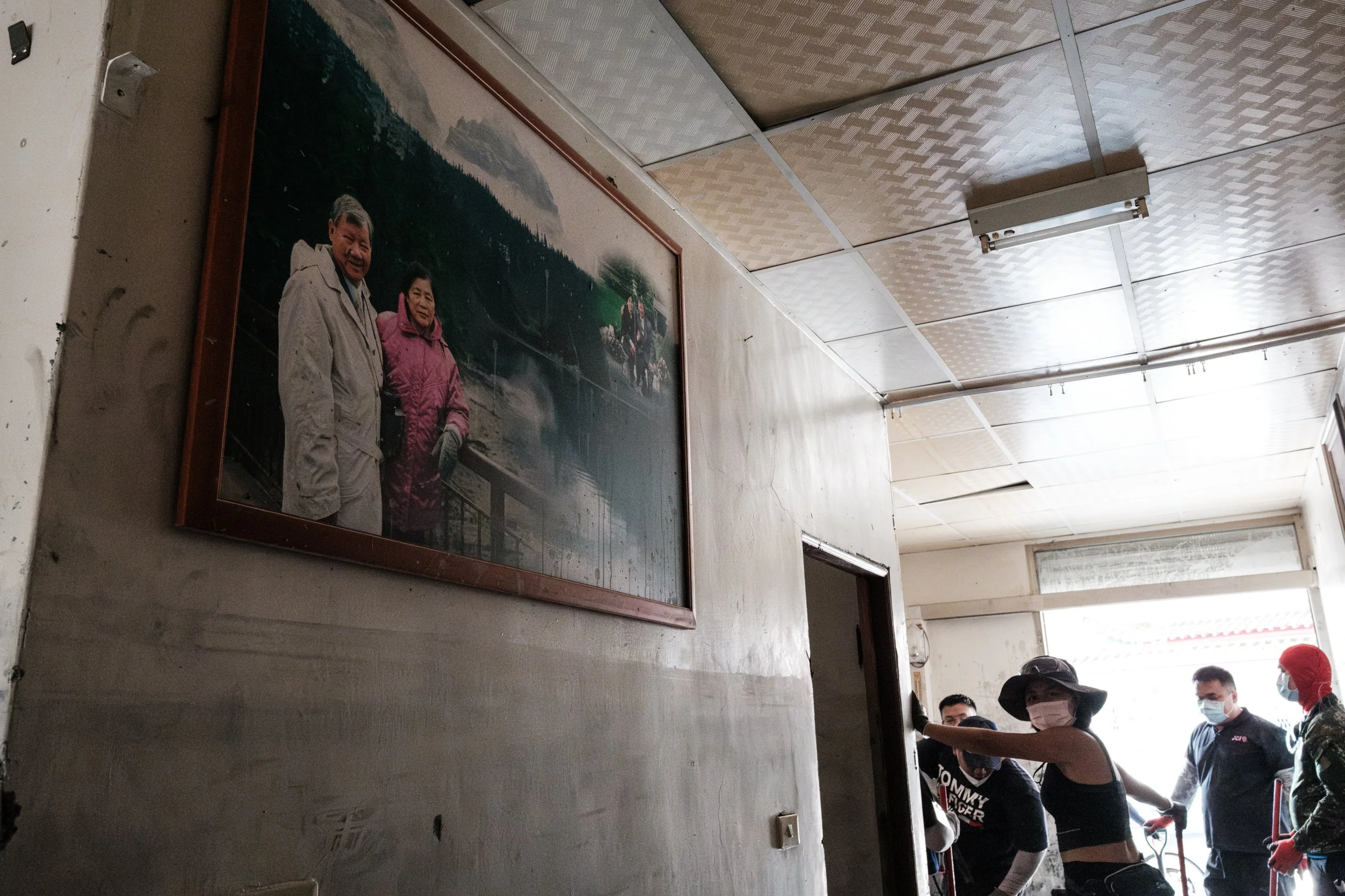 A photo of a couple is on the wall of a home destroyed by water and mud in the 2025 Guangfu flood. A visible water line can be seen just below the photo and volunteers are in the background. 