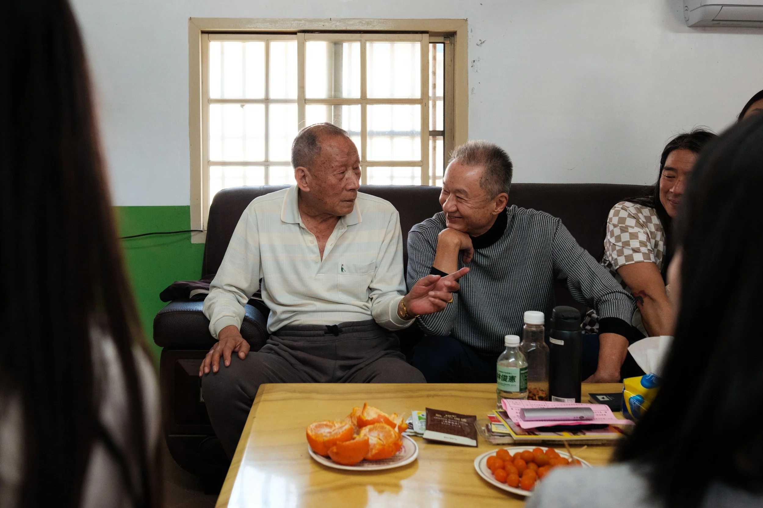 Two older men are talking on a sofa in the middle of a Chinese New Year family gathering in Tainan. One man is smiling as the other one talks and gestures with his hand.