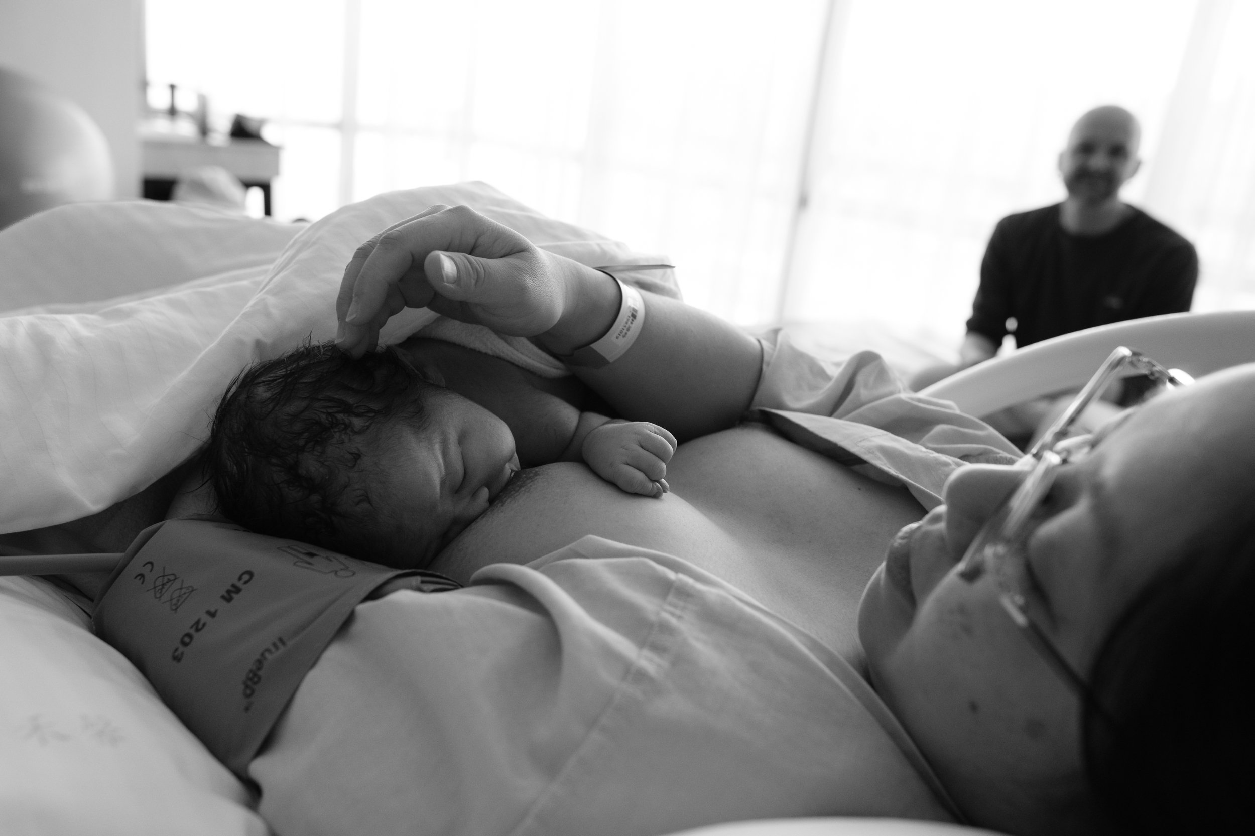 Black and white birth photography image of a newborn resting skin-to-skin on their mother's chest at Dianthus, with the father visible in the background by a sunlit window.