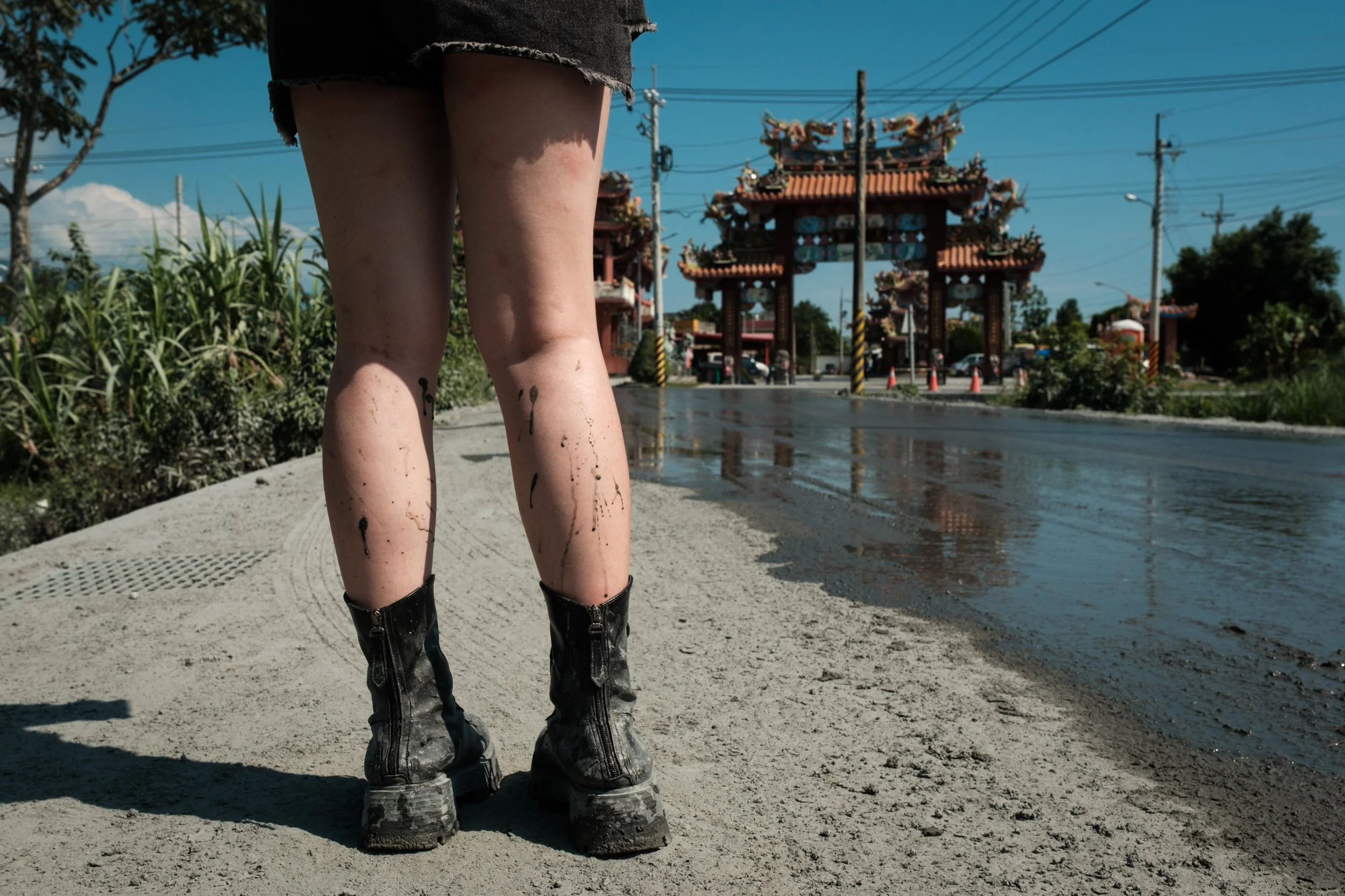 A woman's muddy legs are in the foreground and a temple gate is in the background. She is standing on a muddy street in Guangfu Township in Hualien County. 