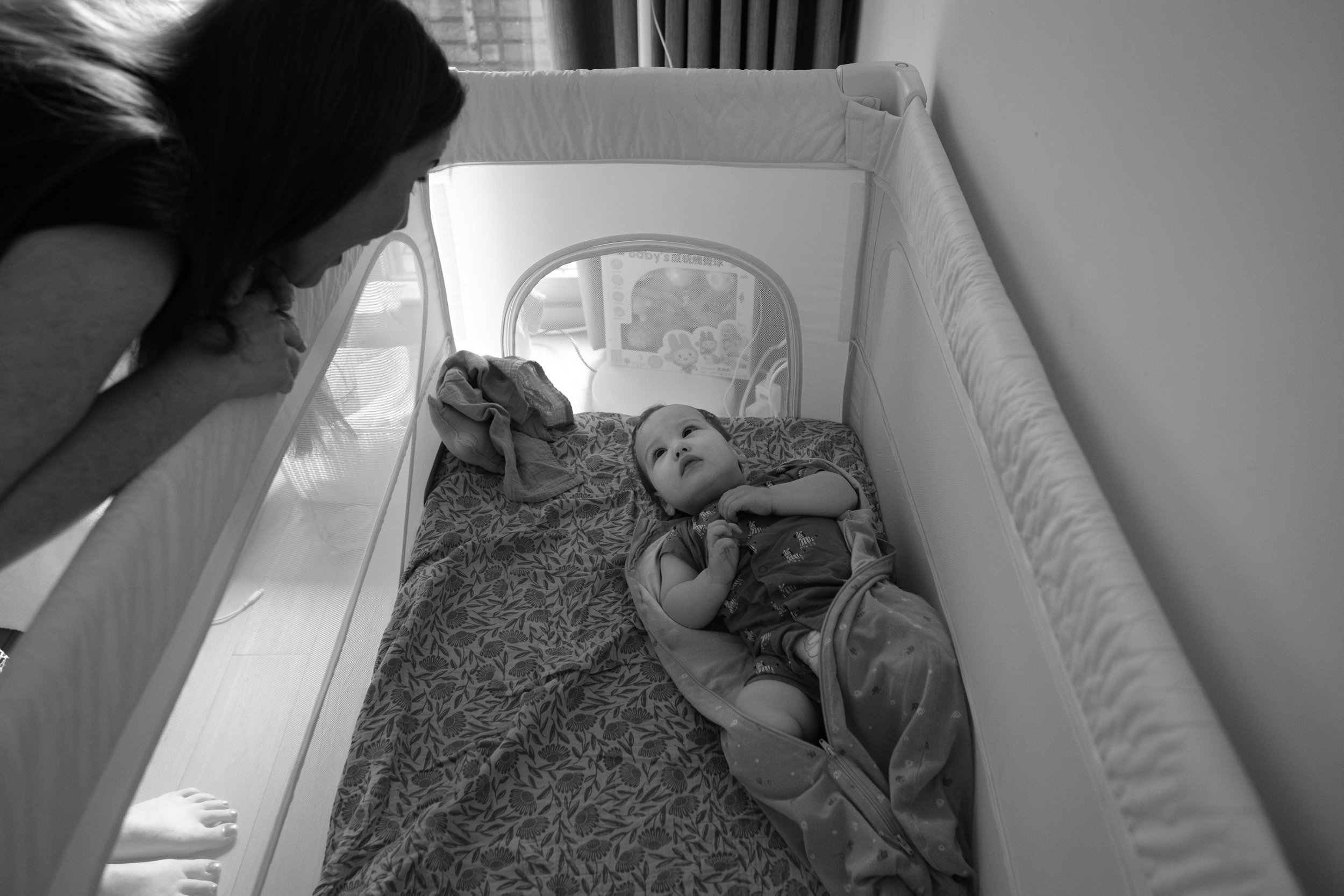A baby is in his crib and looking up at his mother as she leans over and wakes him up from his nap.
