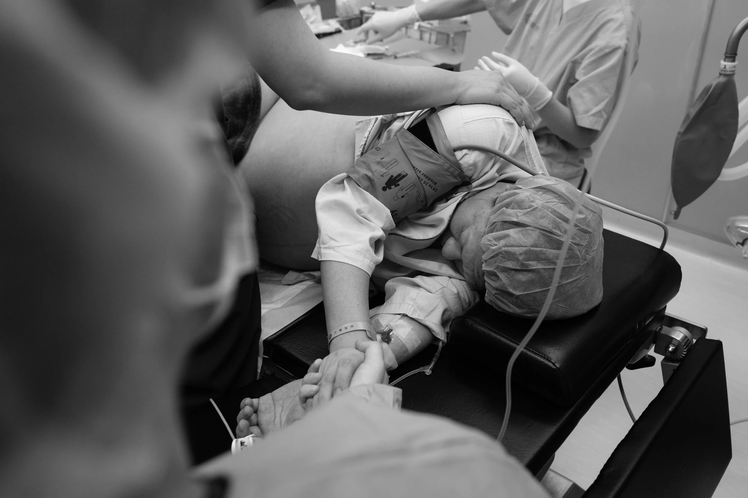 Black and white birth photography image of a mother being prepared for a c-section at Dianthus, medical staff attaching monitoring equipment as someone holds her hand.