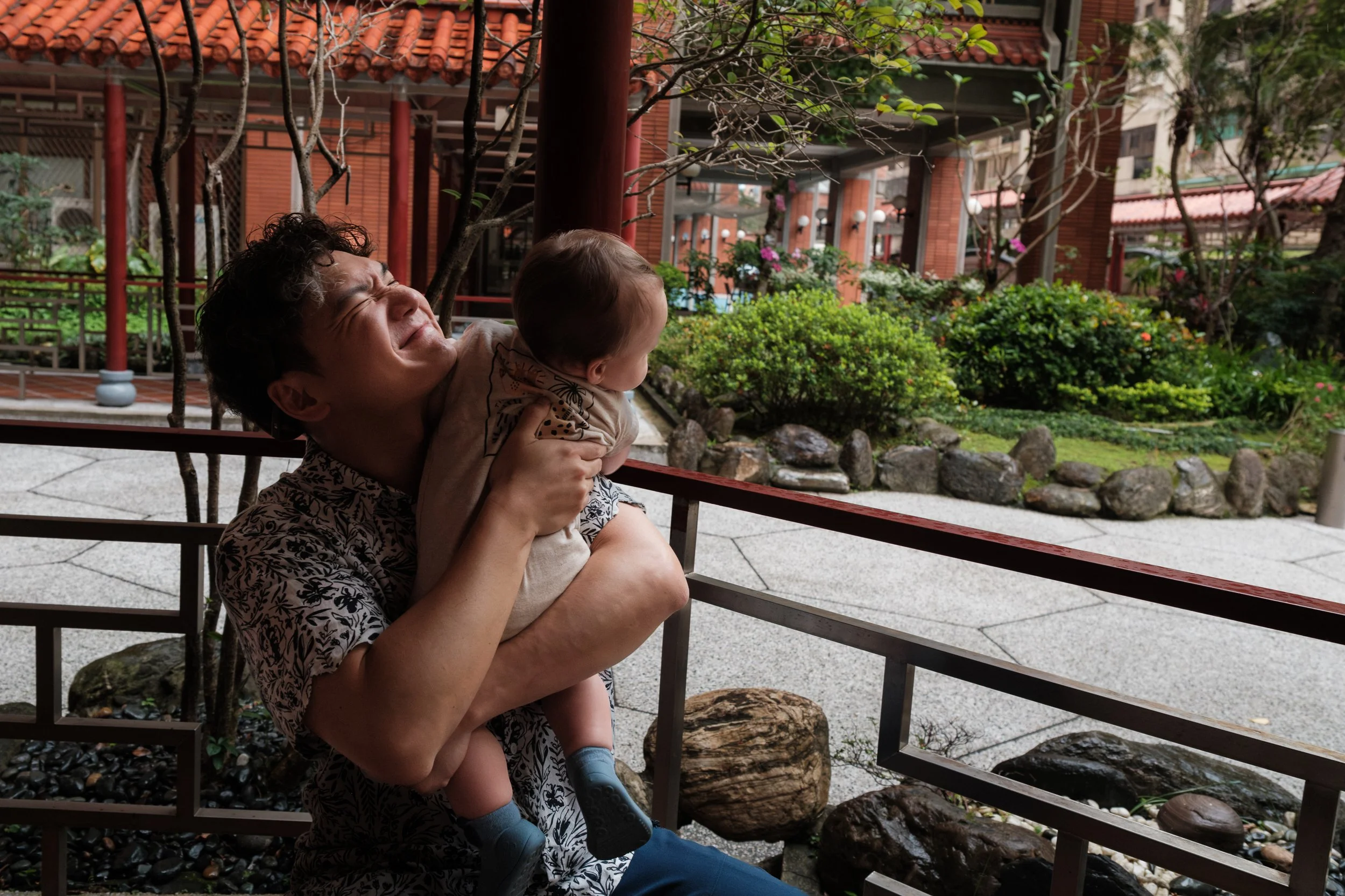 A father is scrunching his face up and smiling as his toddler son grabs his collar. Photographed during an outdoor family photography session in Taipei.
