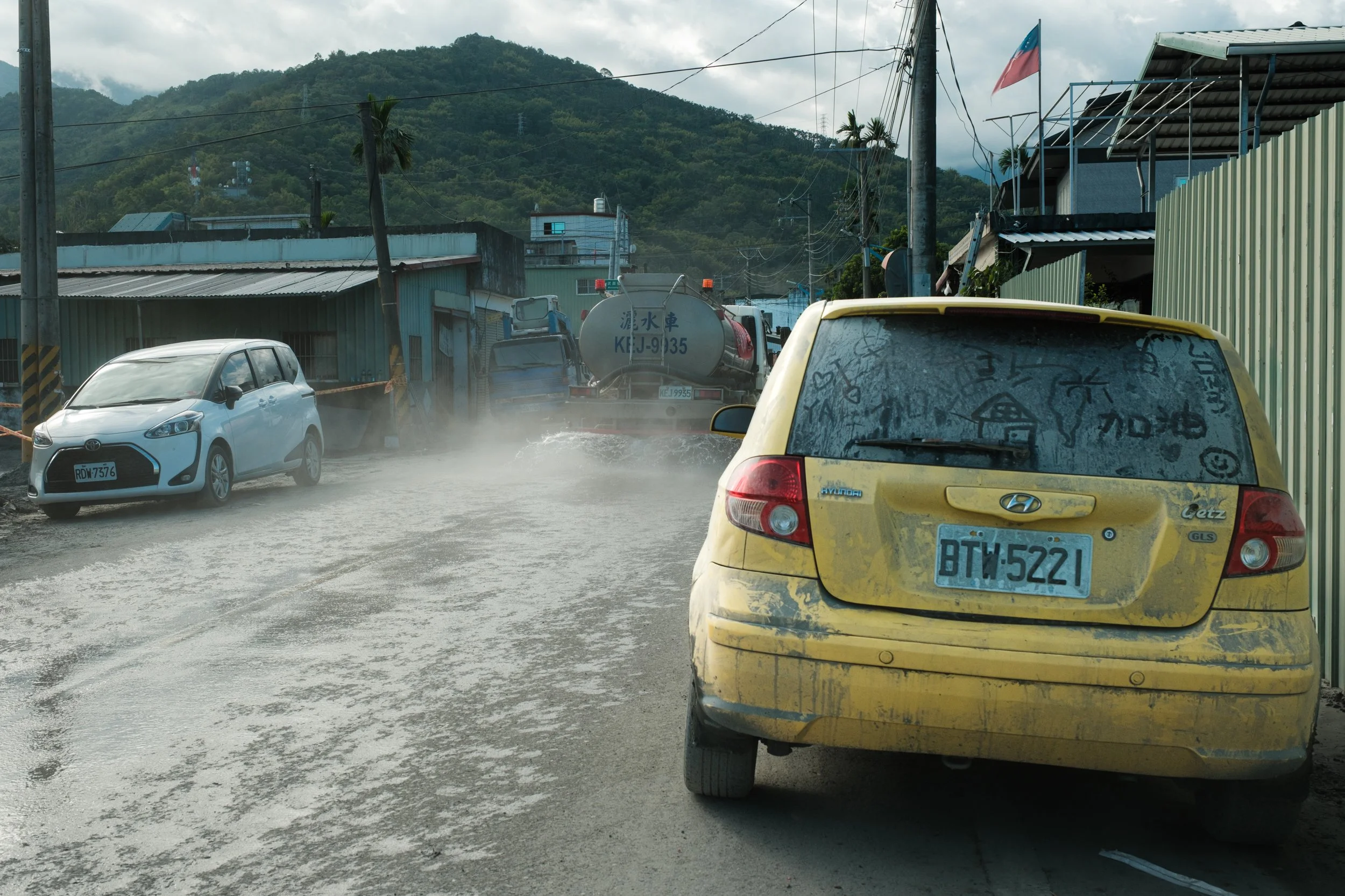 A yellow car is parked on the side of a dusty, muddy road in Guangfu. Messages of encouragement are written in the dirt of the car and a street cleaning car is driving down the road. 