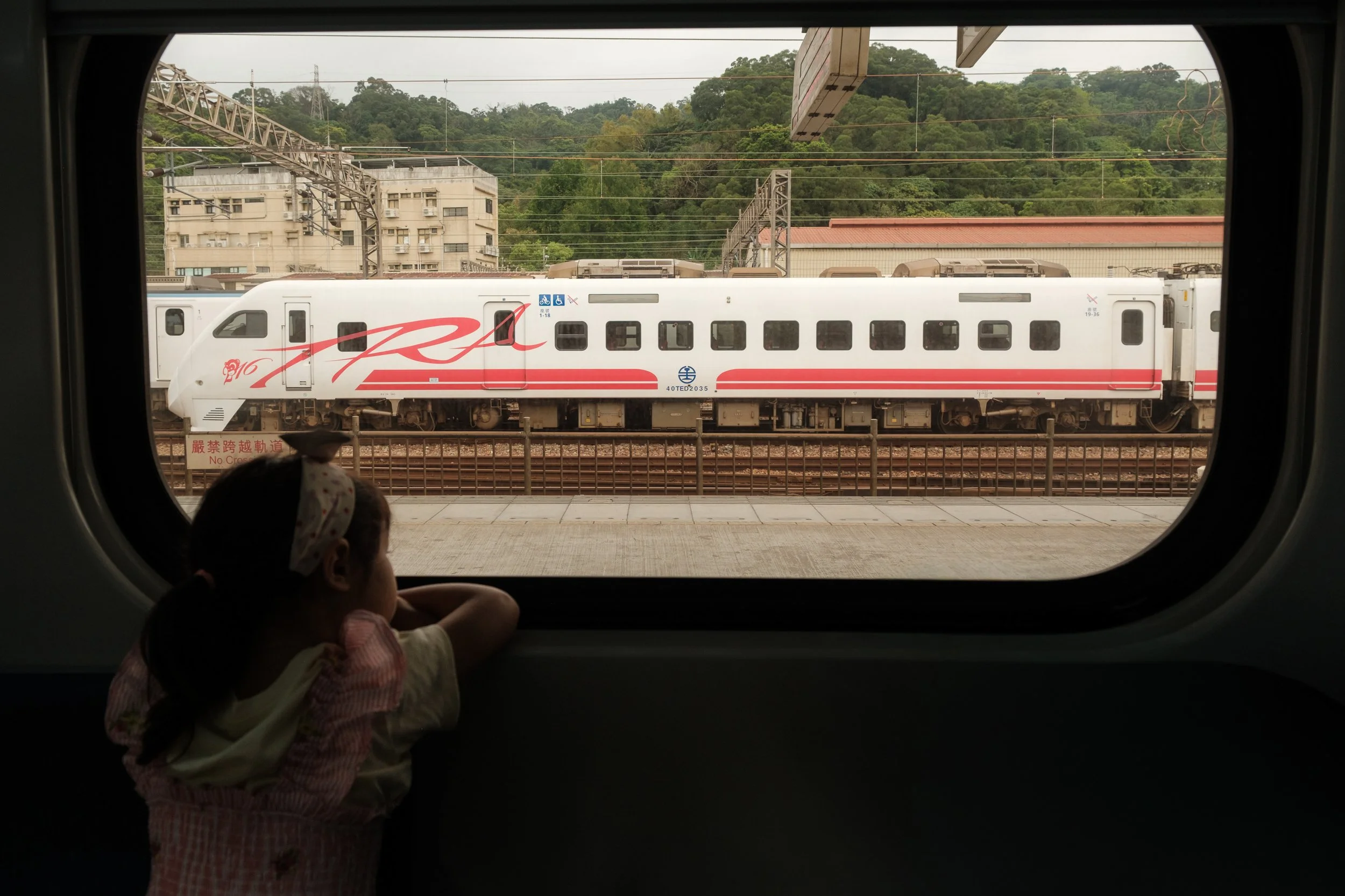 A little girl is looking out the window of a train that is traveling from Taipei to Yingge. Another train can be seen through the window.