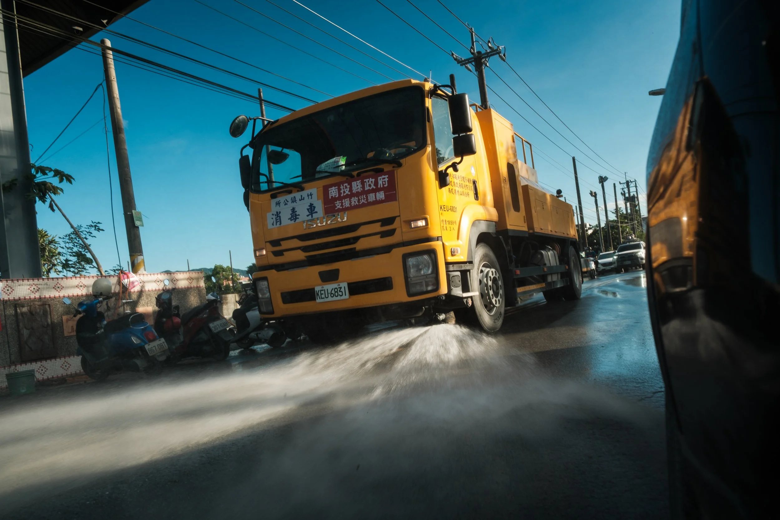 A yellow street-cleaning truck is driving down a muddy street in Guangfu Township and blasting water everywhere in order to clean the street after the flood.