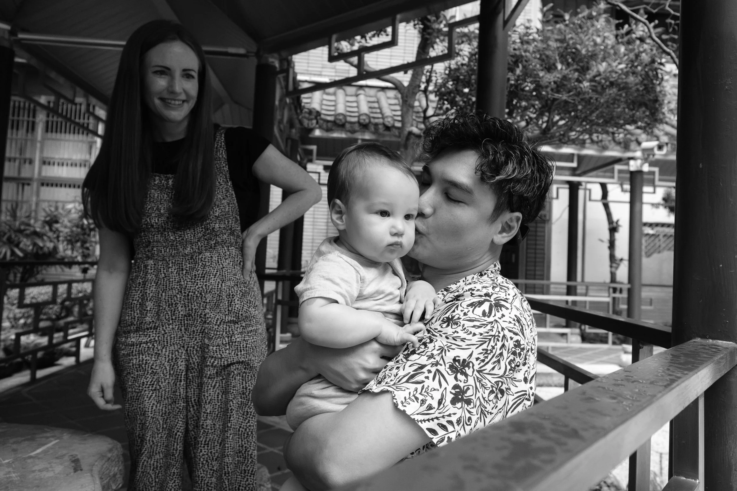 A father is kissing his baby son during a Taipei family photoshoot as his mother watches and smiles in the background.