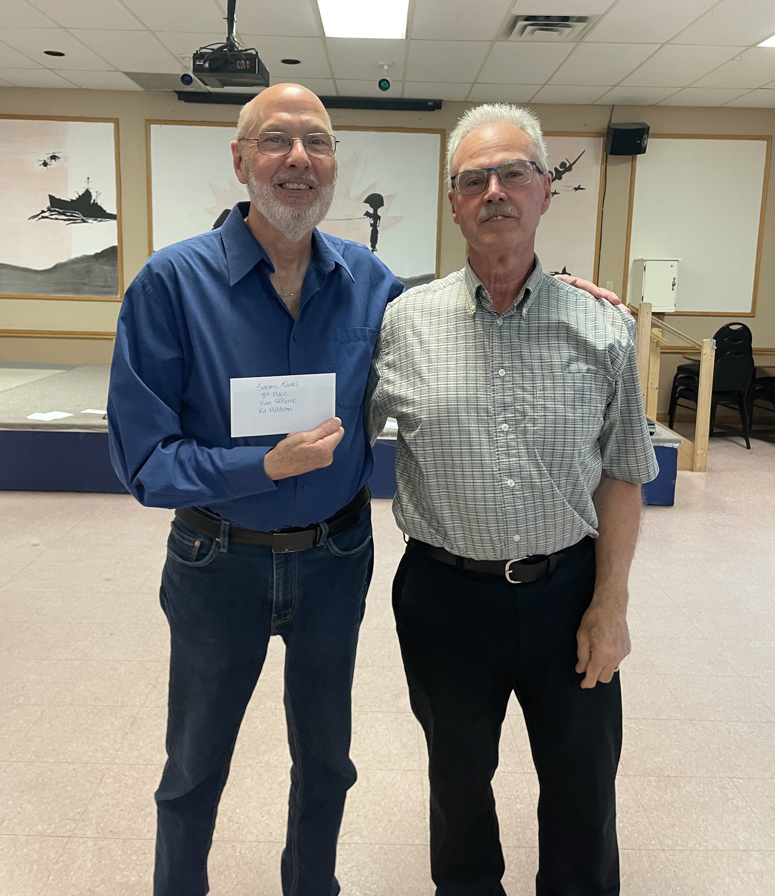 Two elderly men standing together indoors, one holding a note. They are smiling and wearing glasses. The background has painted walls with military or war-themed artwork.