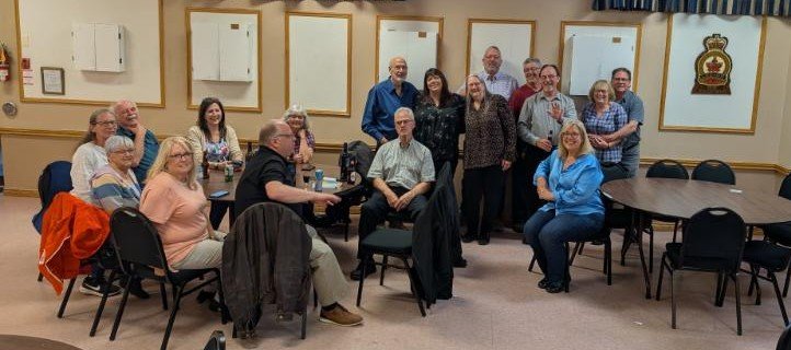 Group of adults gathered in a conference room, some seated around tables and others standing, posing for a photo.
