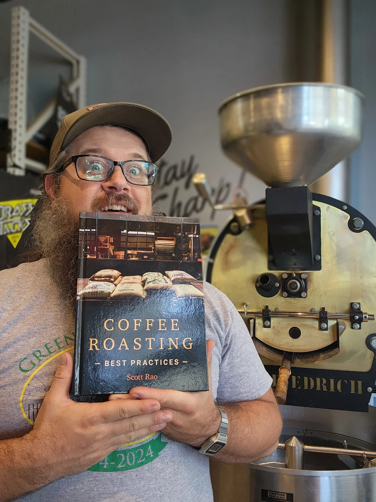 Justin, Roasting and Wholesale Director at Brass Tacks Coffee Co., in roastery holding coffee roasting book next to roasting machine