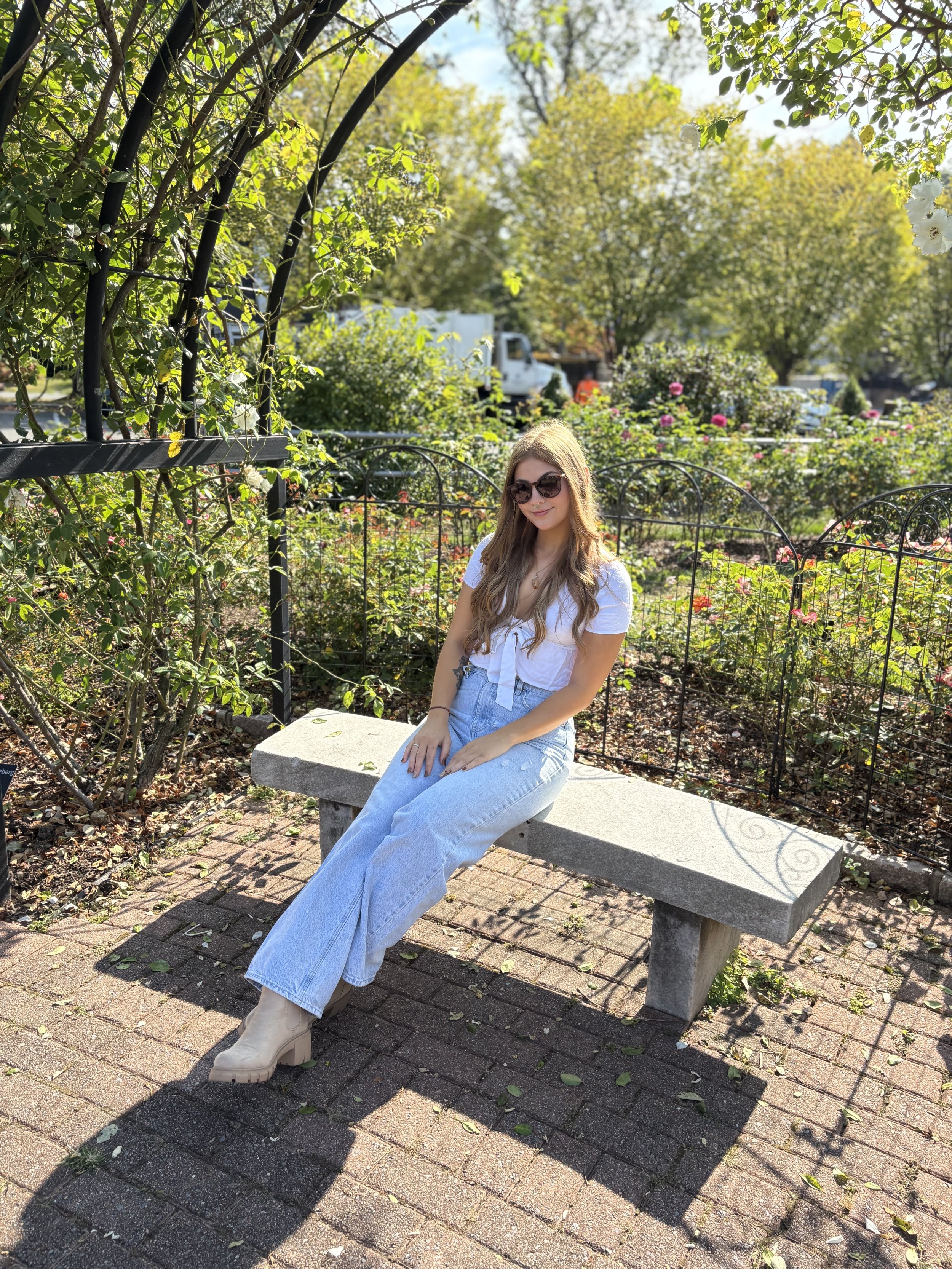 Gianna Tabbitas, Southside manager, wearing sunglasses, a white top, light blue jeans, and beige boots sitting on a stone bench in a park surrounded by greenery and blooming flowers, with trees and a truck in the background.