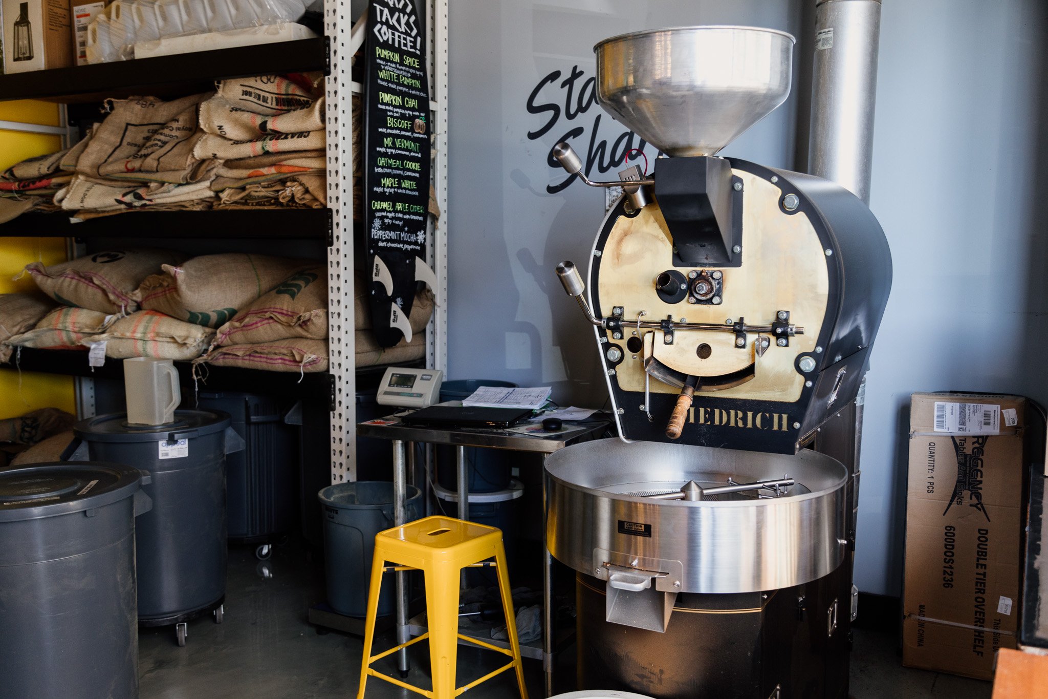 A coffee roasting machine in a storage room with shelves holding burlap sacks, a black chalkboard sign listing different coffee flavors, and various containers and boxes.