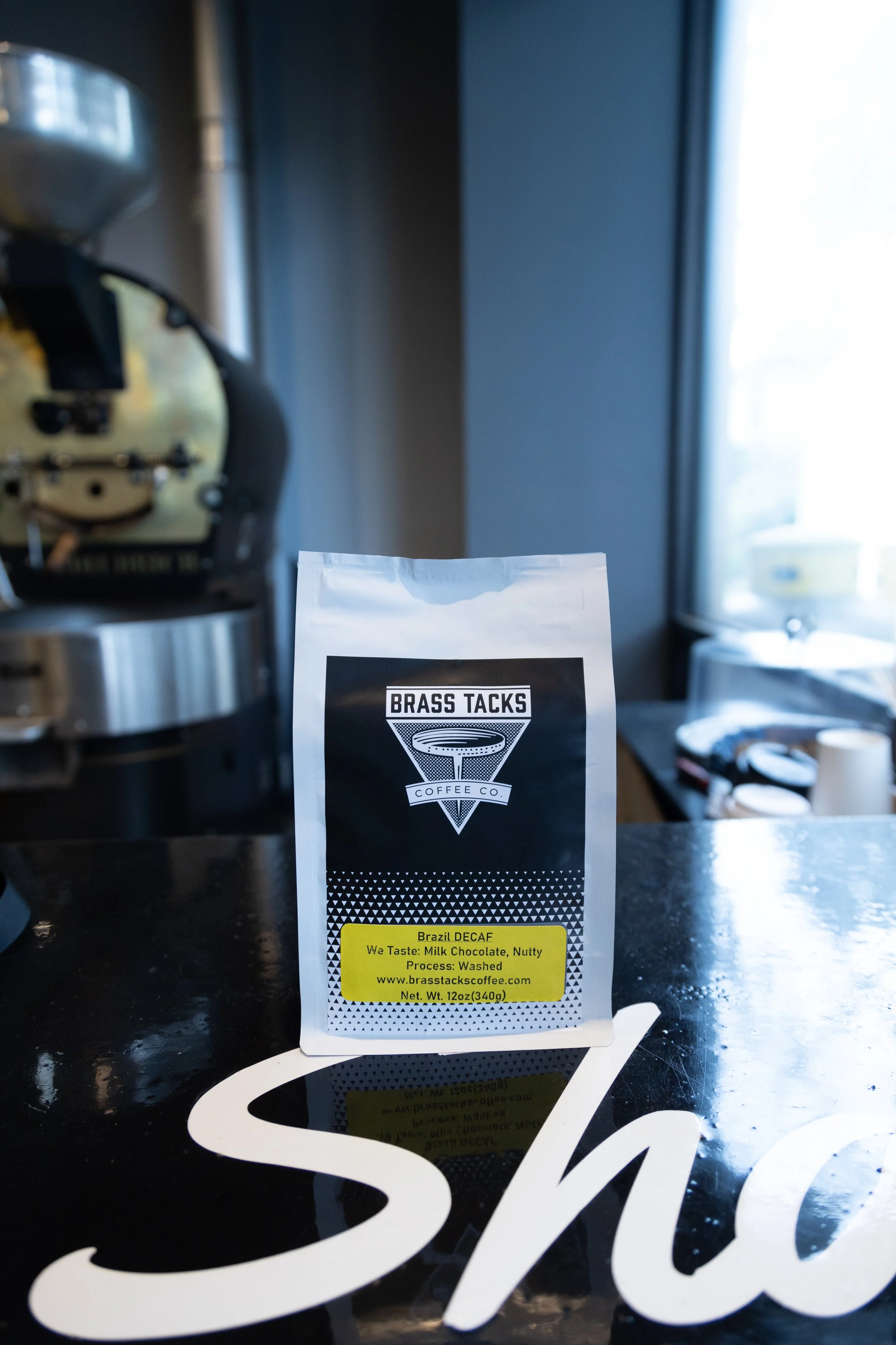 Coffee bag labeled 'Brass Tacks Coffee Co.' with a black and white geometric design and yellow text indicating decaffeinated Brazil coffee, placed on a reflective black surface in a coffee shop setting with a coffee machine and window in the background.