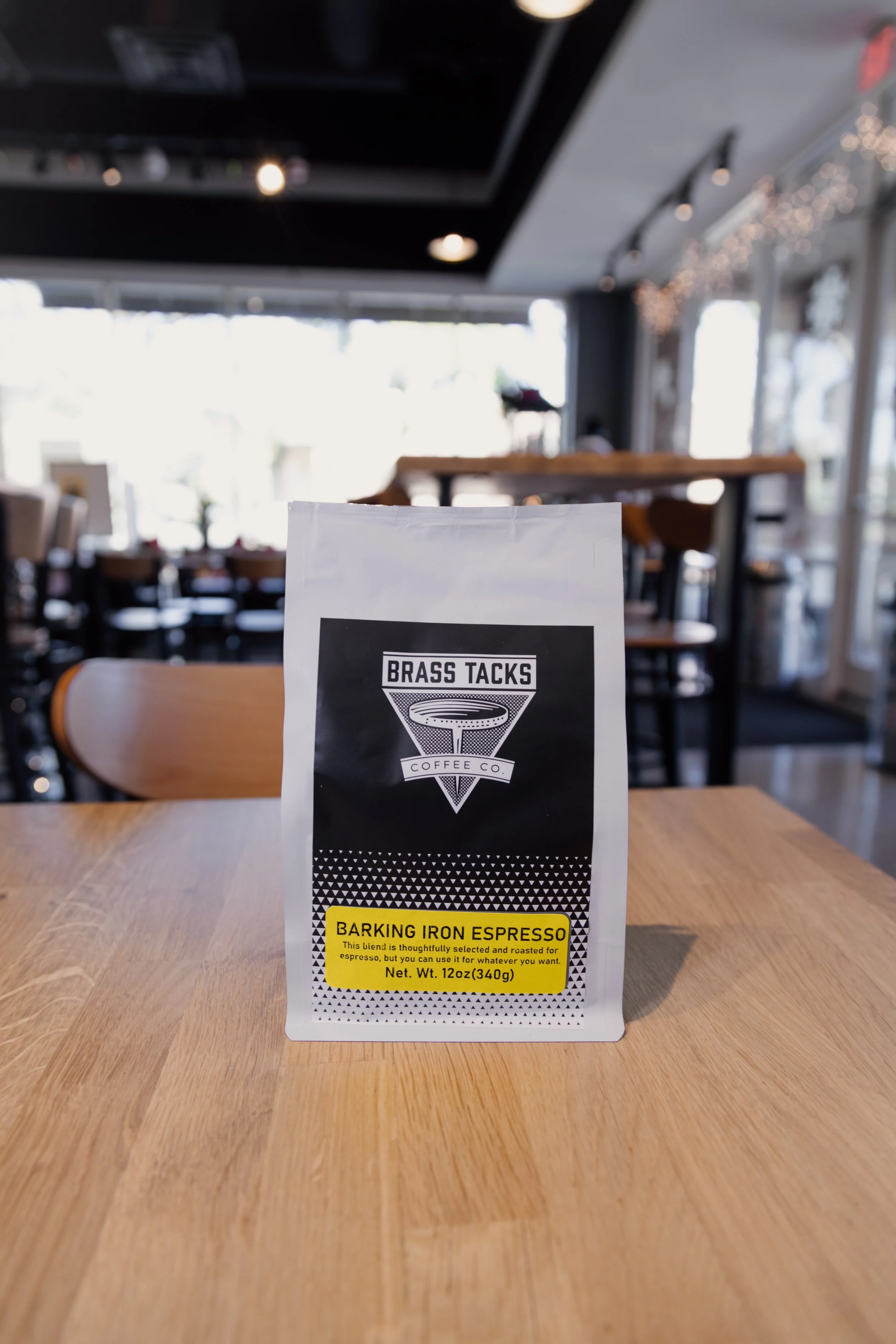A package of Brass Tacks Coffee Co.'s Barking Iron Espresso coffee beans on a wooden table inside a bright cafe with large windows and string lights.