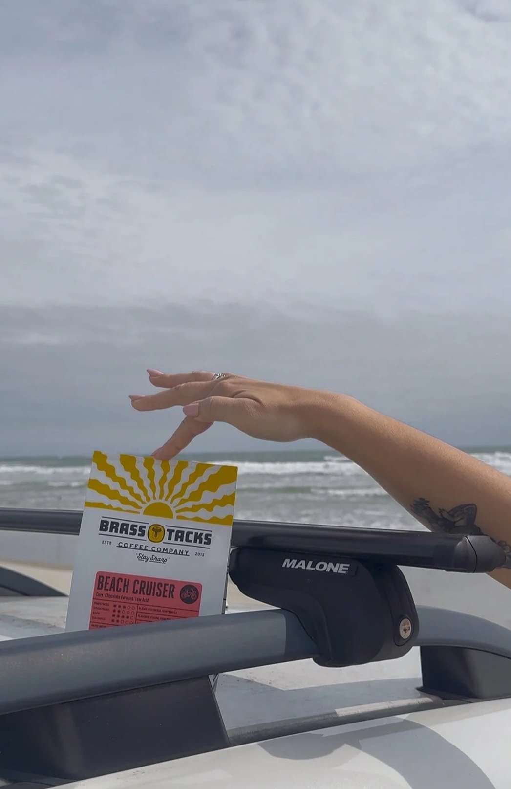A person's hand holding a Brass Tacks Coffee Company loyalty card featuring a sun and wave design, with a tattoo visible on the wrist, against a beach background with cloudy sky and ocean waves.