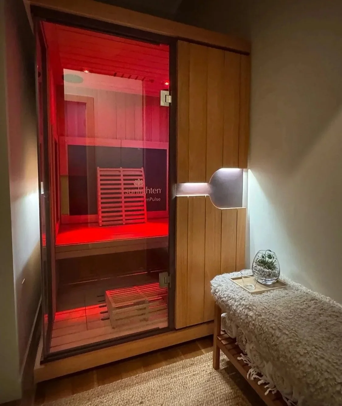 Indoor infrared sauna room with red lighting, wooden interior, glass door, and a small wooden bench inside. Next to it, a cozy area with a textured blanket, glass containers, and a small plant in a glass vase.