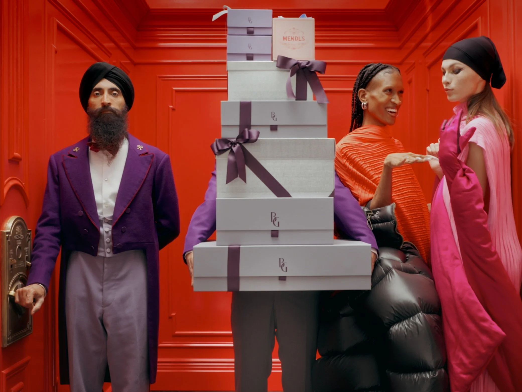 People in colorful outfits in front of a red wall, one person is holding a stack of boxes tied with purple ribbons.