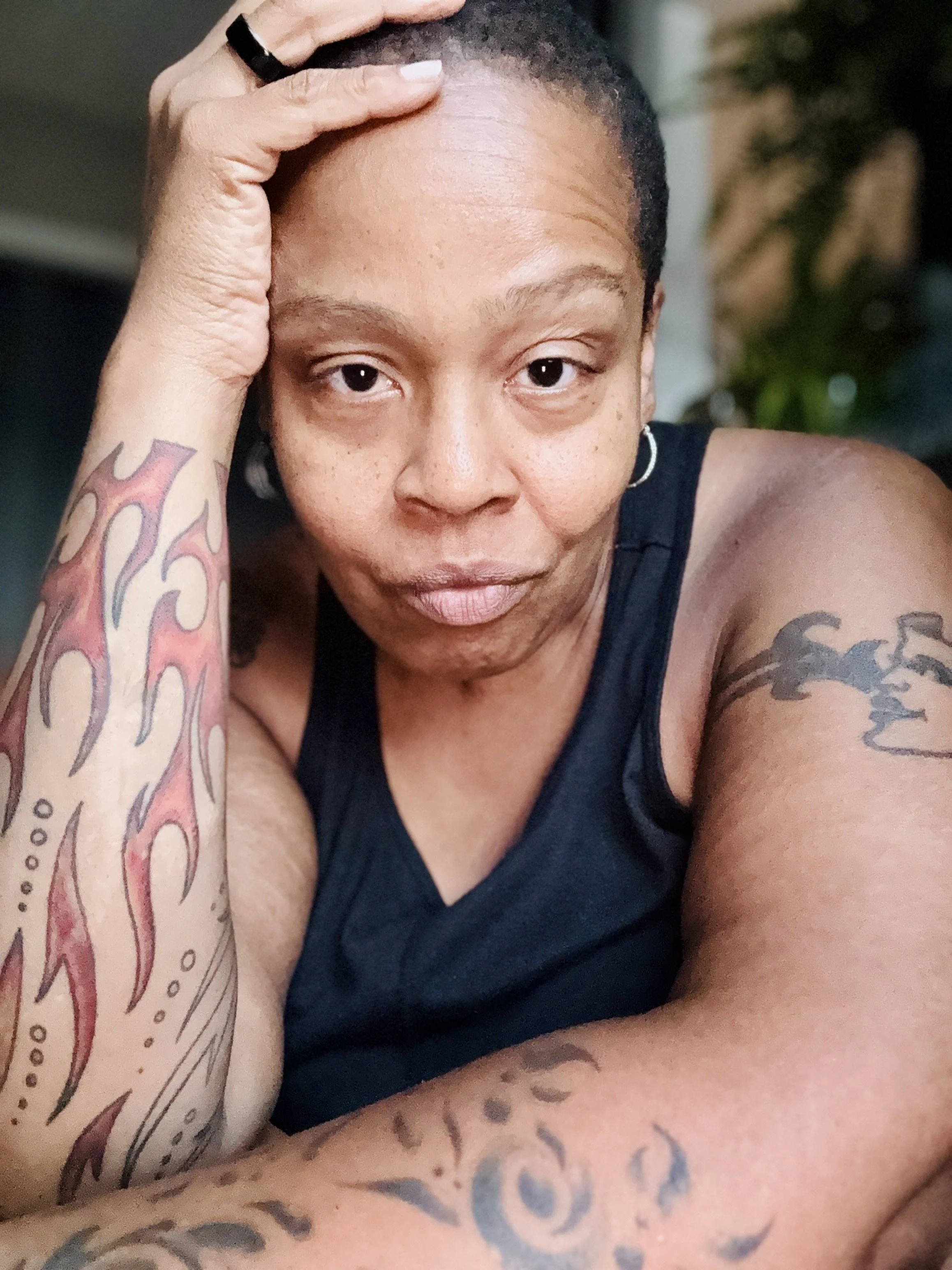 Erica DeLaine Bradshaw, tattoos on her arms, wearing a black sleeveless top, resting her head on her hand.