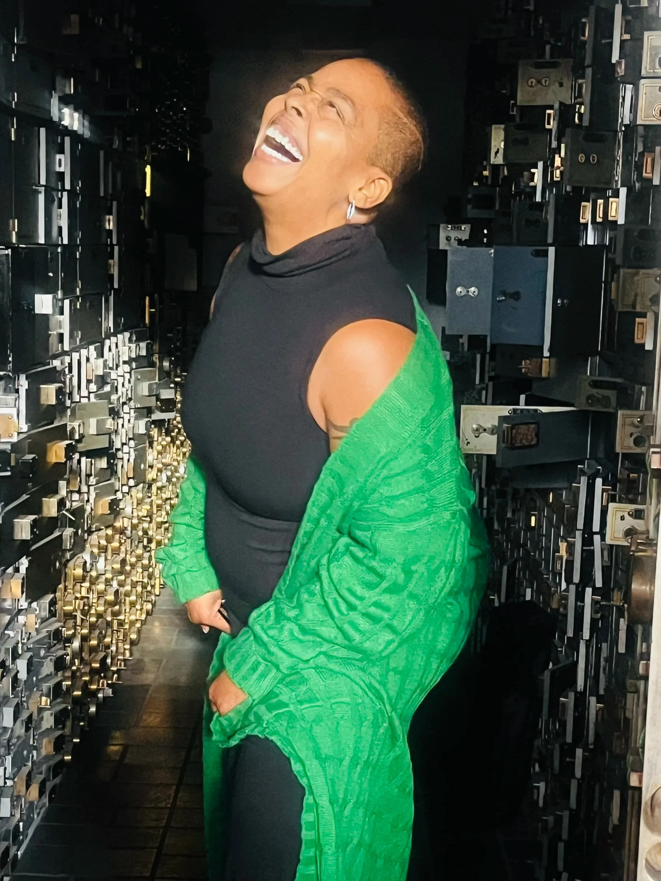 Erica DeLaine Bradshaw  laughing joyfully in a room with walls lined with dark safes or vaults.