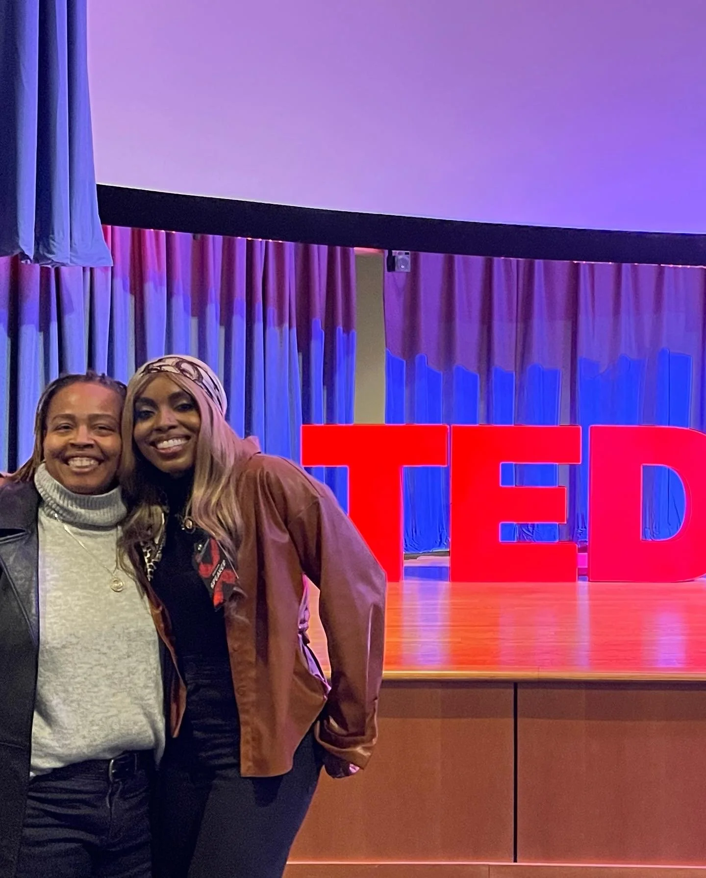 Two women smiling and standing in front of a large red TED sign on a stage with purple curtains and lighting.